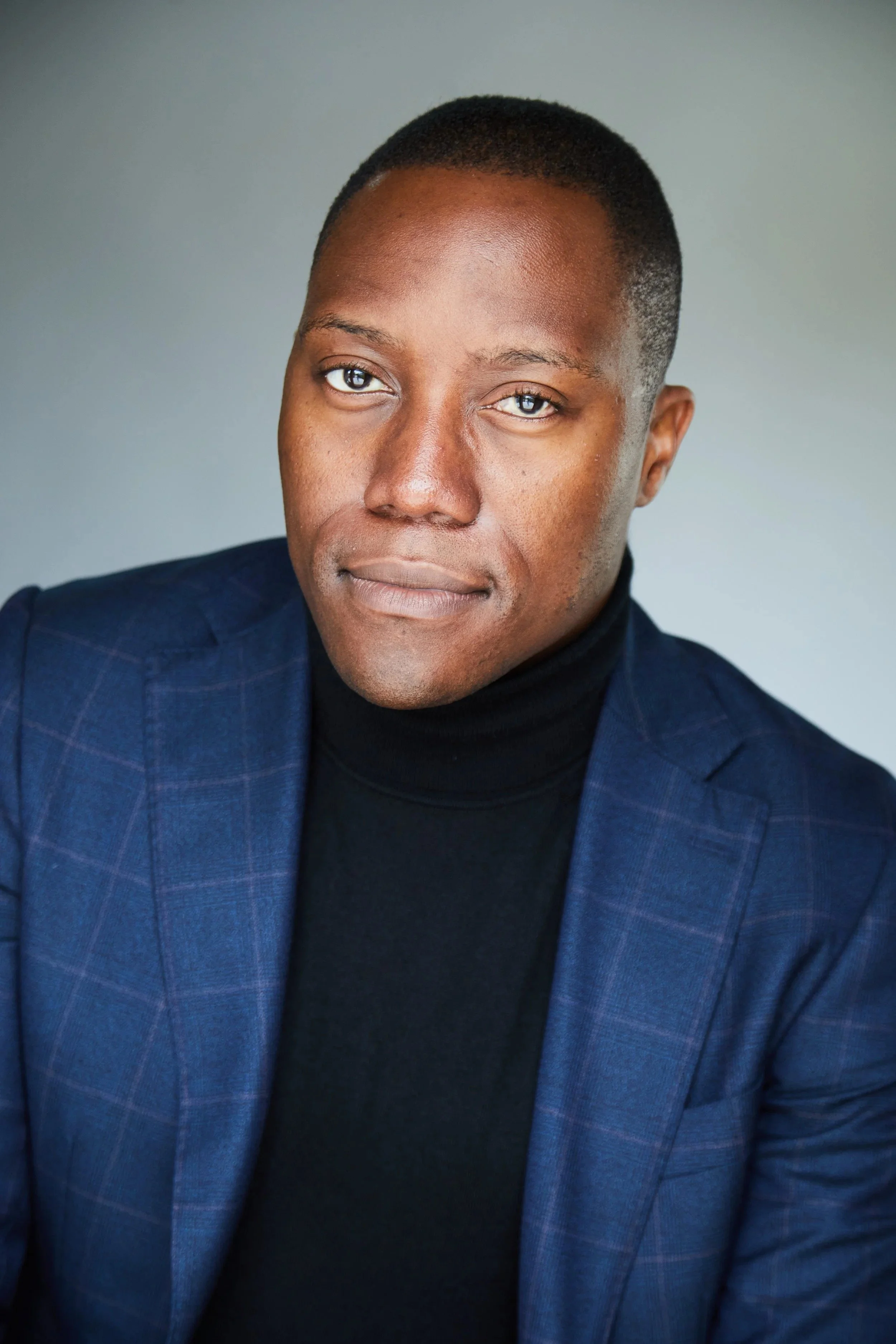 A portrait of a young man with short black hair, wearing a black turtleneck and a blue blazer, looking directly at the camera with a neutral expression.