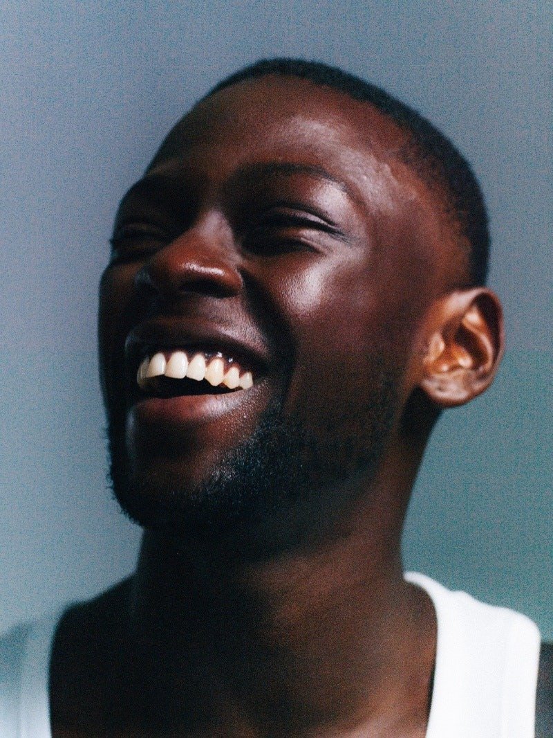 Close-up of a young black man smiling broadly, showing white teeth, with a short haircut and beard, wearing a white shirt against a plain background.