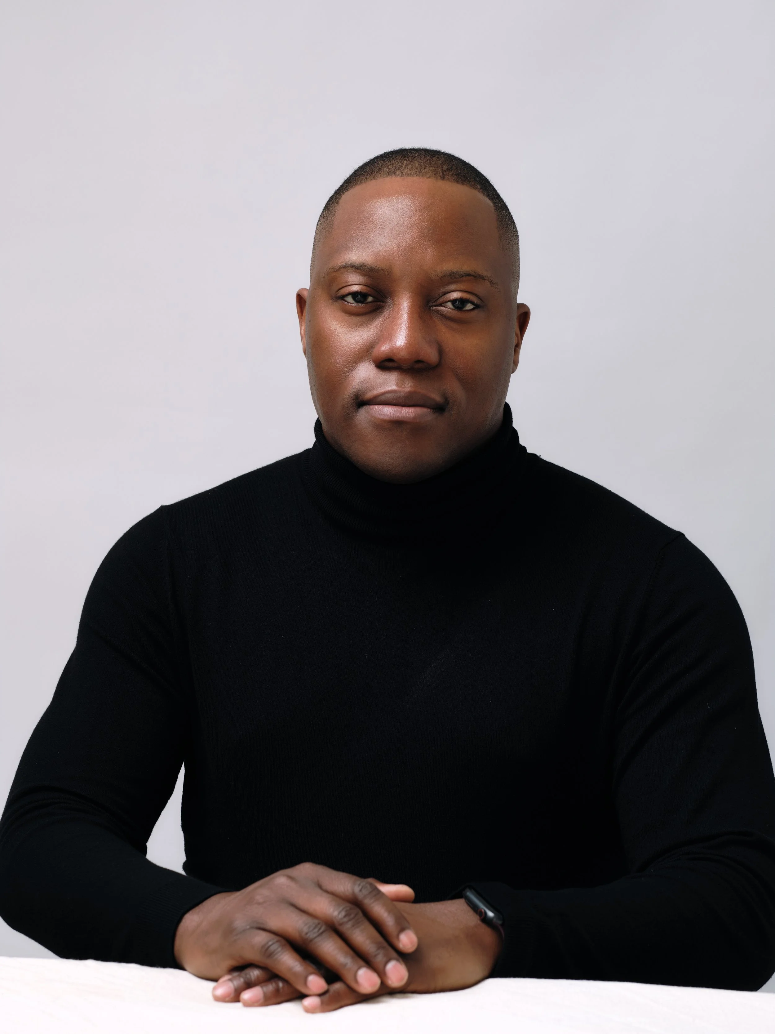 Portrait of Dr Xavier Mulenga, Melbourne psychiatrist, wearing a black turtleneck, seated with hands on a white surface, against a plain white background.
