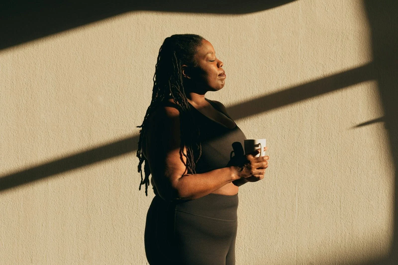 A woman with dreadlocks holding a white mug, standing against a textured beige wall with shadows cast across her face and body.
