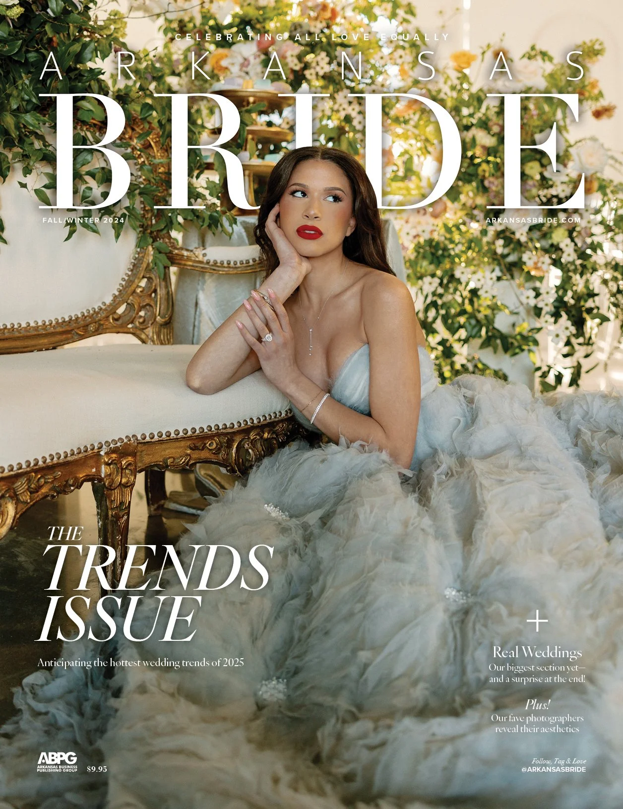 A woman in a strapless wedding gown sitting on the floor with her arm resting on a vintage cream-colored sofa with gold accents, surrounded by lush flowers and greenery, on the cover of Arkansas Bride magazine's Fall/Winter 2024 issue.