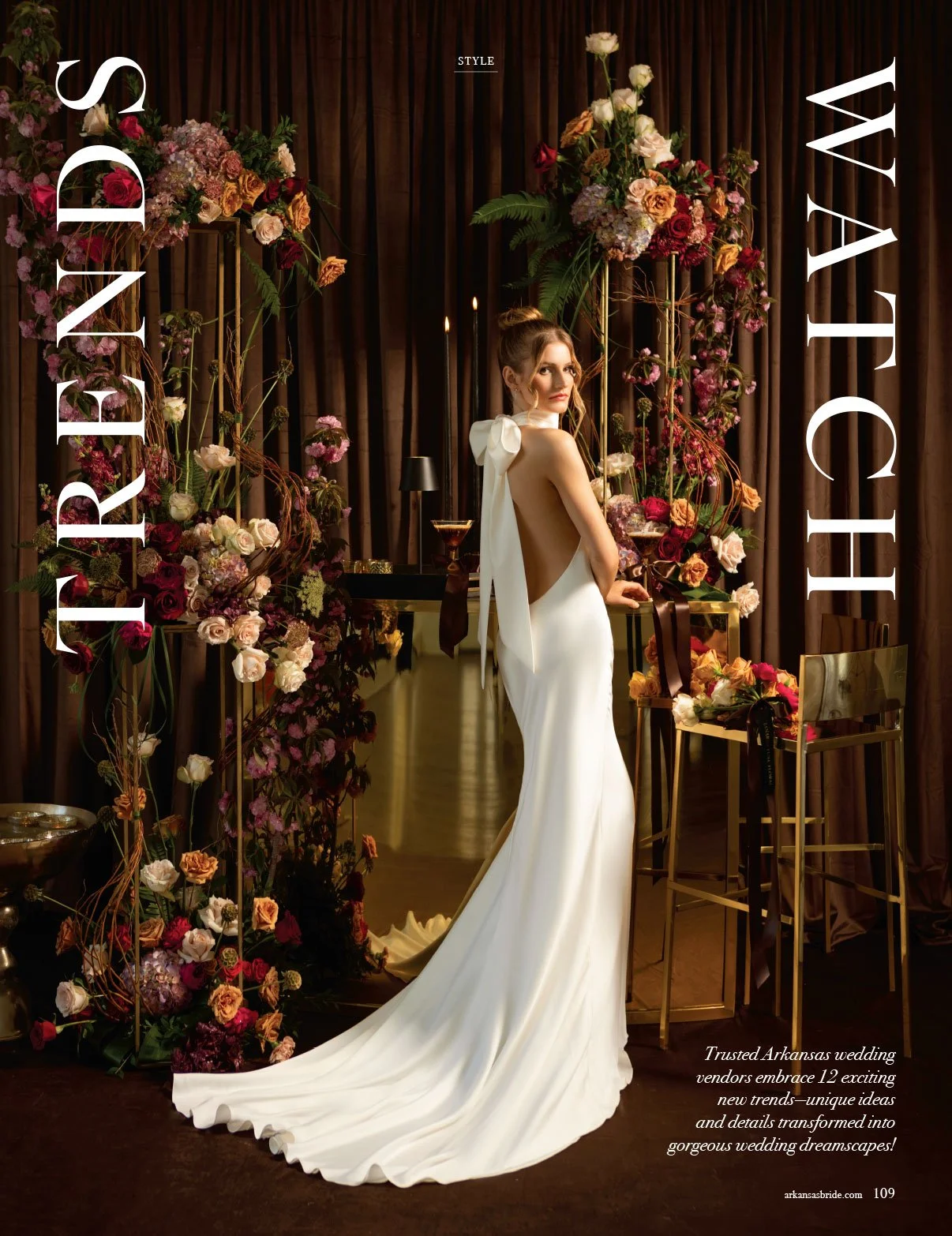 A woman in a backless white wedding gown with a long train, standing among floral arrangements and gold furniture, with dark curtains in the background.