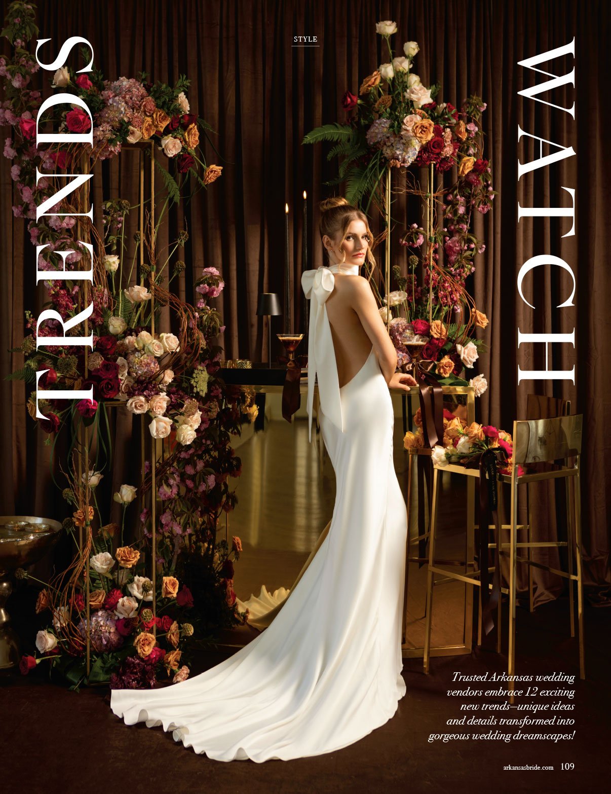 A fashion model in a backless white gown with a long train standing among floral arrangements and gold furniture at a wedding-themed display.