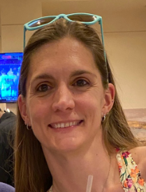 Close-up of a woman smiling with glasses resting on her head, indoors, with a TV screen in the background.