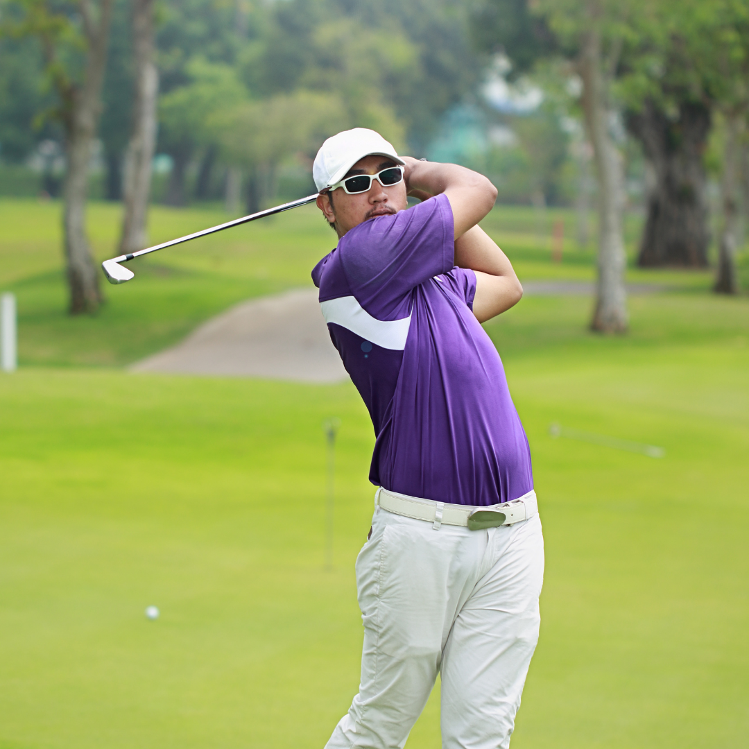 A golfer in purple shirt and white pants swings a golf club on a lush green golf course with trees in the background.