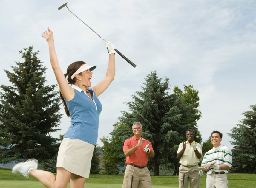 Woman in blue shirt and white skirt holding golf club high on golf course, celebrating with three men in the background.