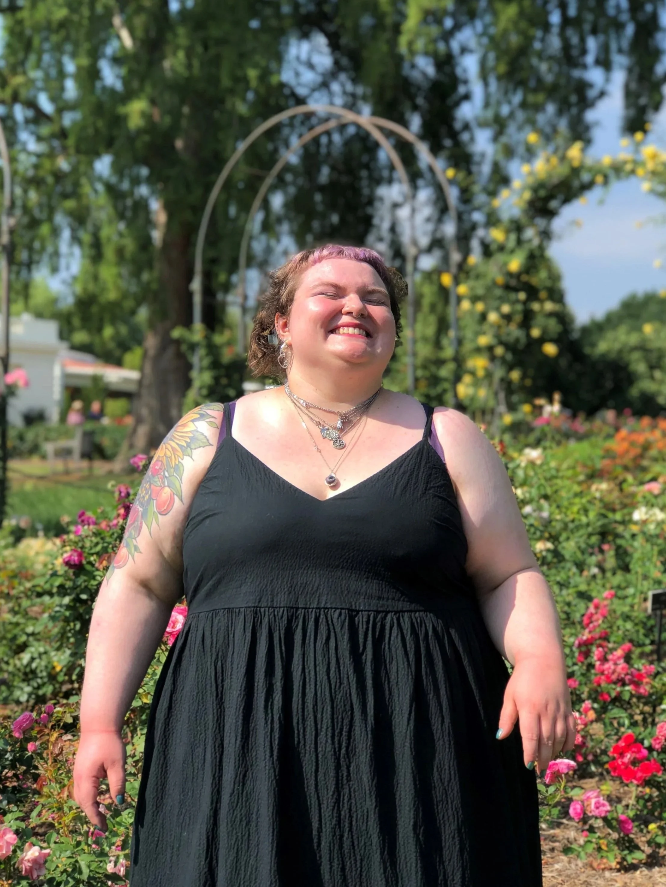 A woman with short hair smiling with eyes closed, wearing a black dress, standing in a garden filled with colorful flowers and greenery.