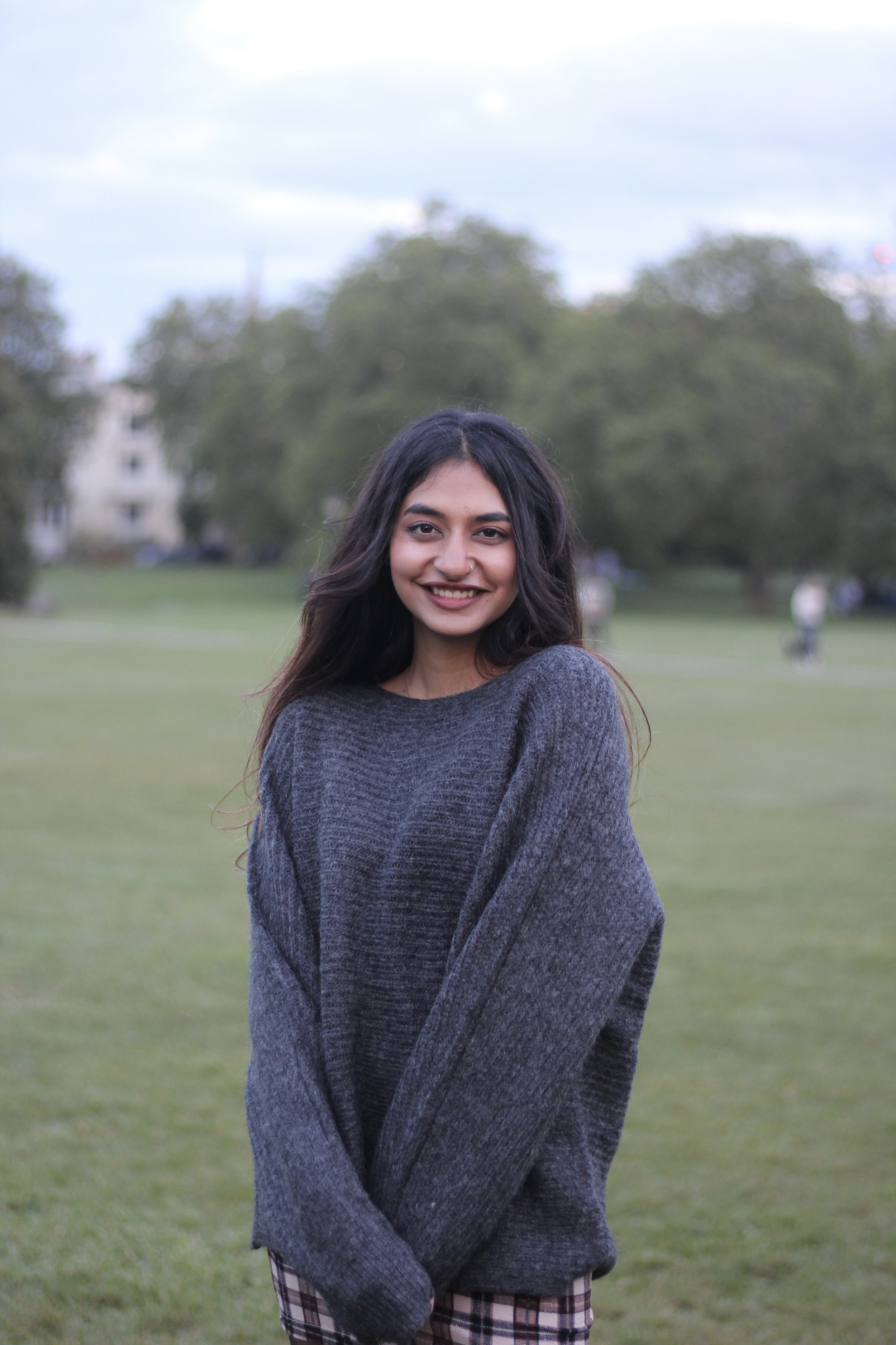 A young woman standing outdoors on a grassy field, smiling at the camera, wearing a dark gray sweater and checkered pants, with trees and a few people in the background.