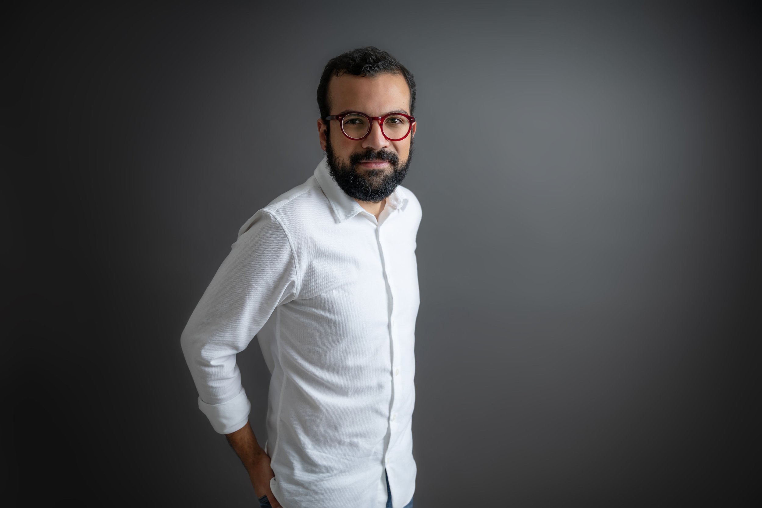 Hombre con barba, gafas rojas y camisa blanca posando de lado en un fondo gris oscuro.