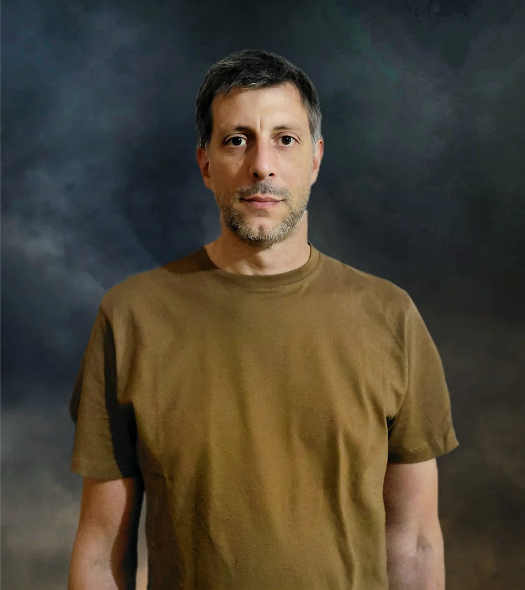 Hombre con barba y camiseta marrón, de fondo oscuro nebuloso.