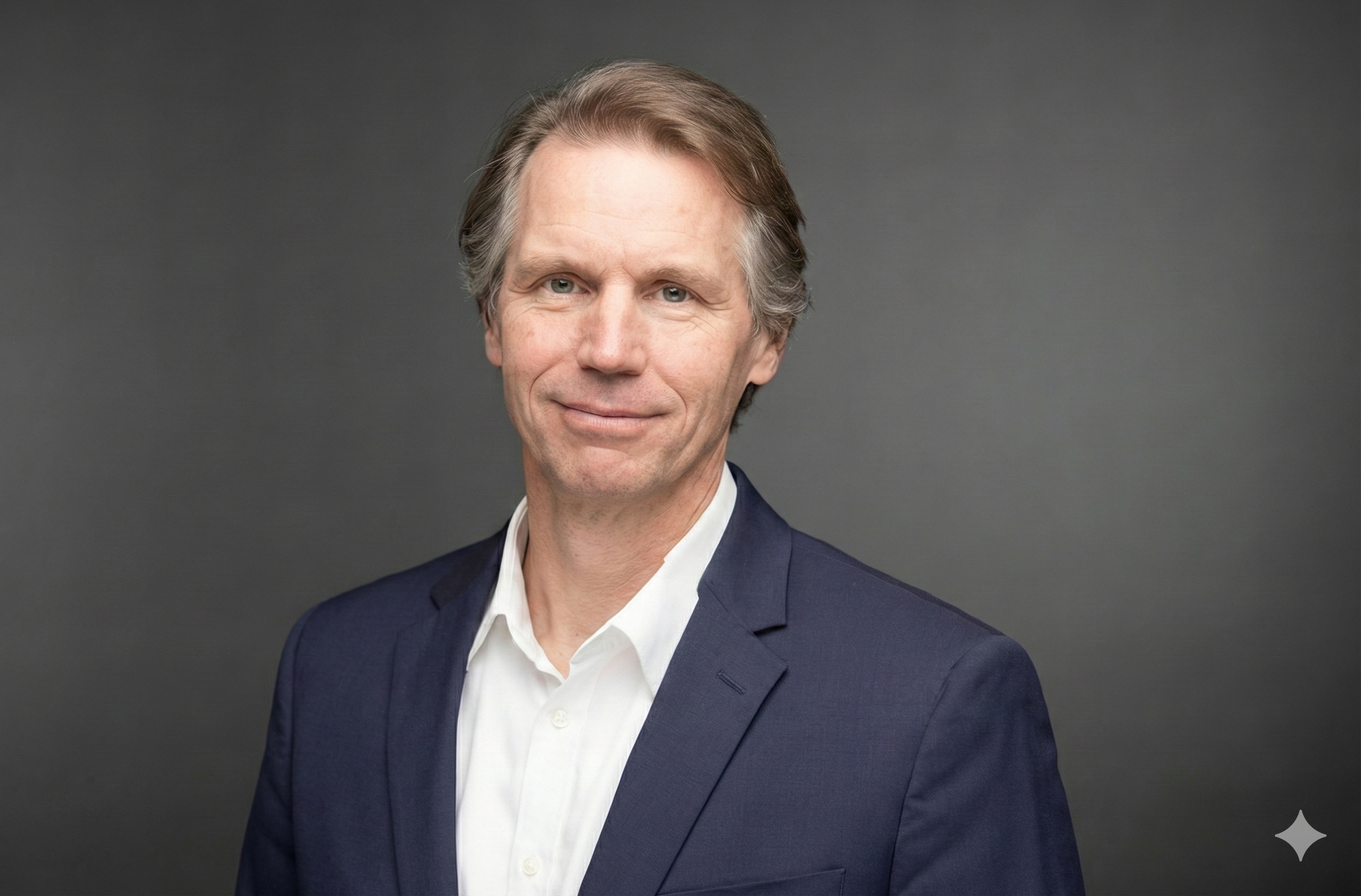 A professional headshot of a middle-aged man with light brown or blonde hair, dressed in a navy suit and white shirt, against a dark gray background.