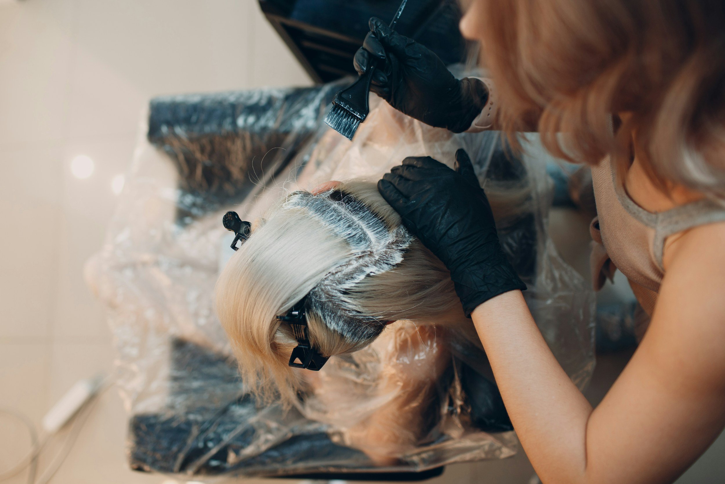 A hairdresser applying a hair dye or bleach treatment to a woman's blonde hair, wearing black gloves and using a brush, with hair sectioned and clipped.