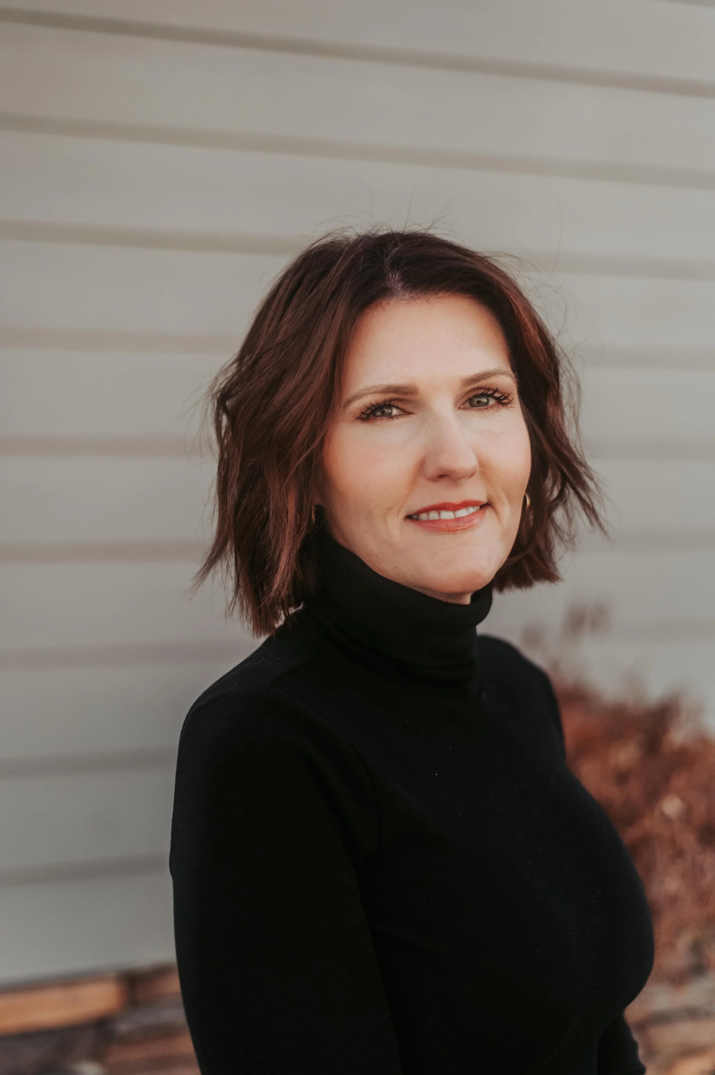 A woman with shoulder-length auburn hair wearing a black turtleneck sweater, standing outdoors in front of a light-colored building with horizontal siding.