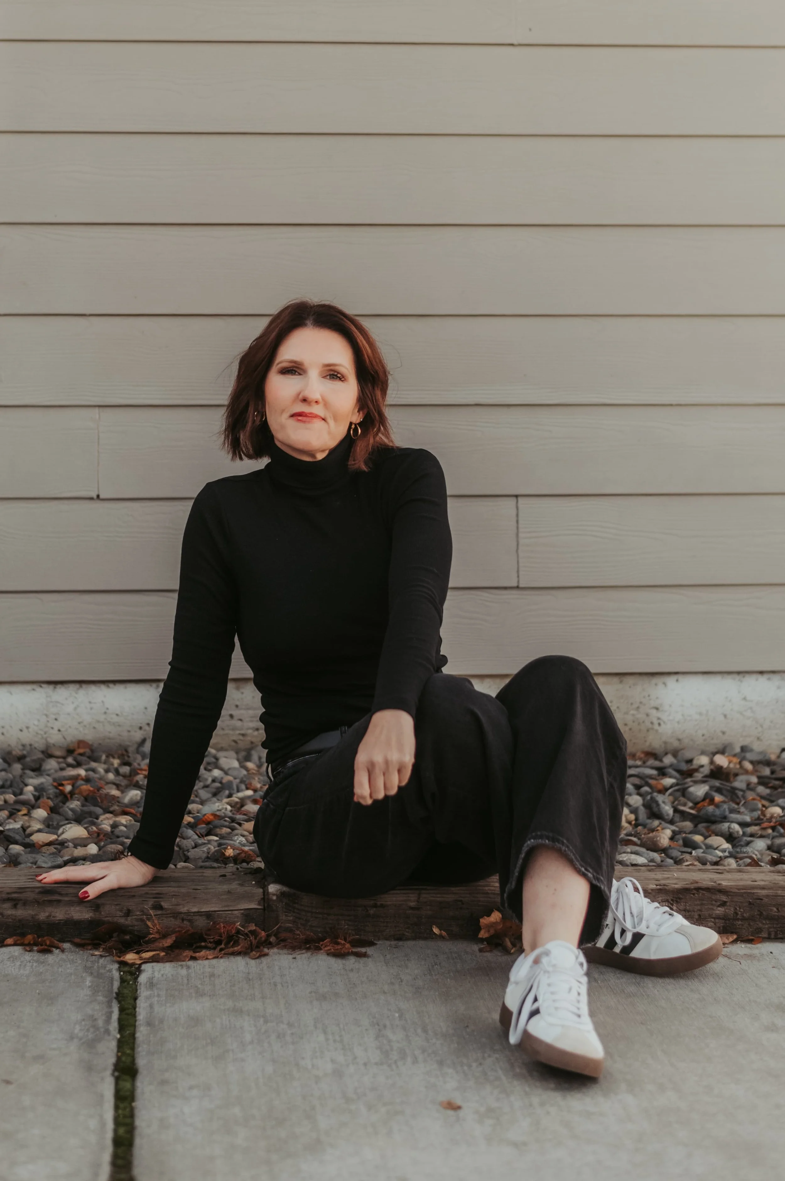 A woman with shoulder-length dark brown hair, wearing a black turtleneck, black pants, and white sneakers, sitting on a wooden curb in front of a gray wooden wall, with rocks and fallen leaves around her.