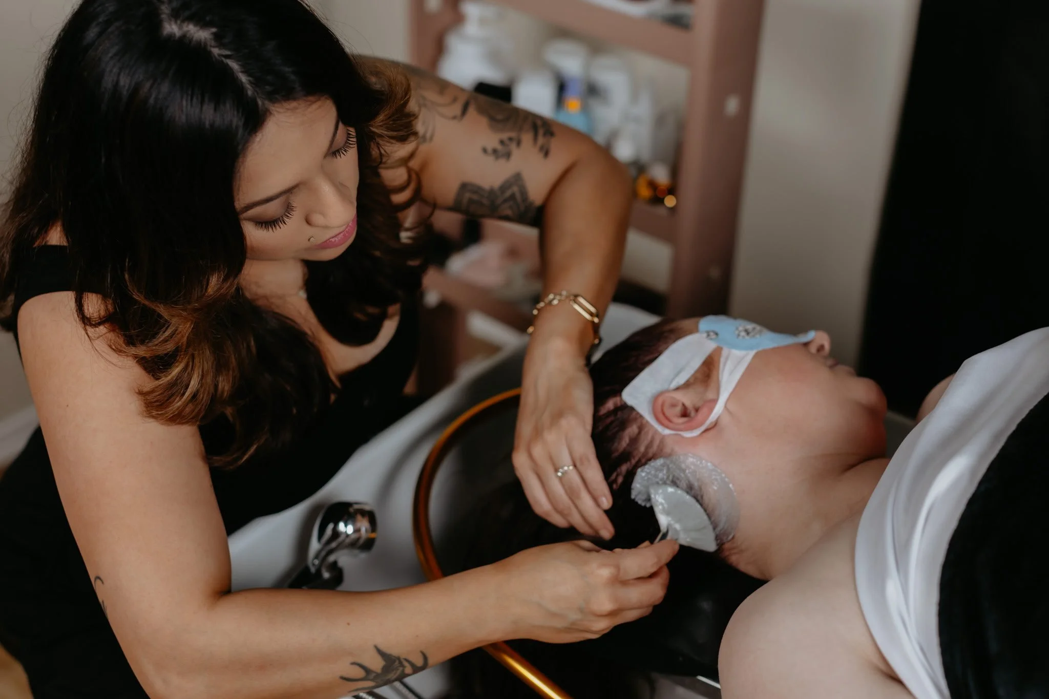 A hairstylist with tattoos and dark hair washing a man's hair in a salon sink while he lies back with a towel on his face.
