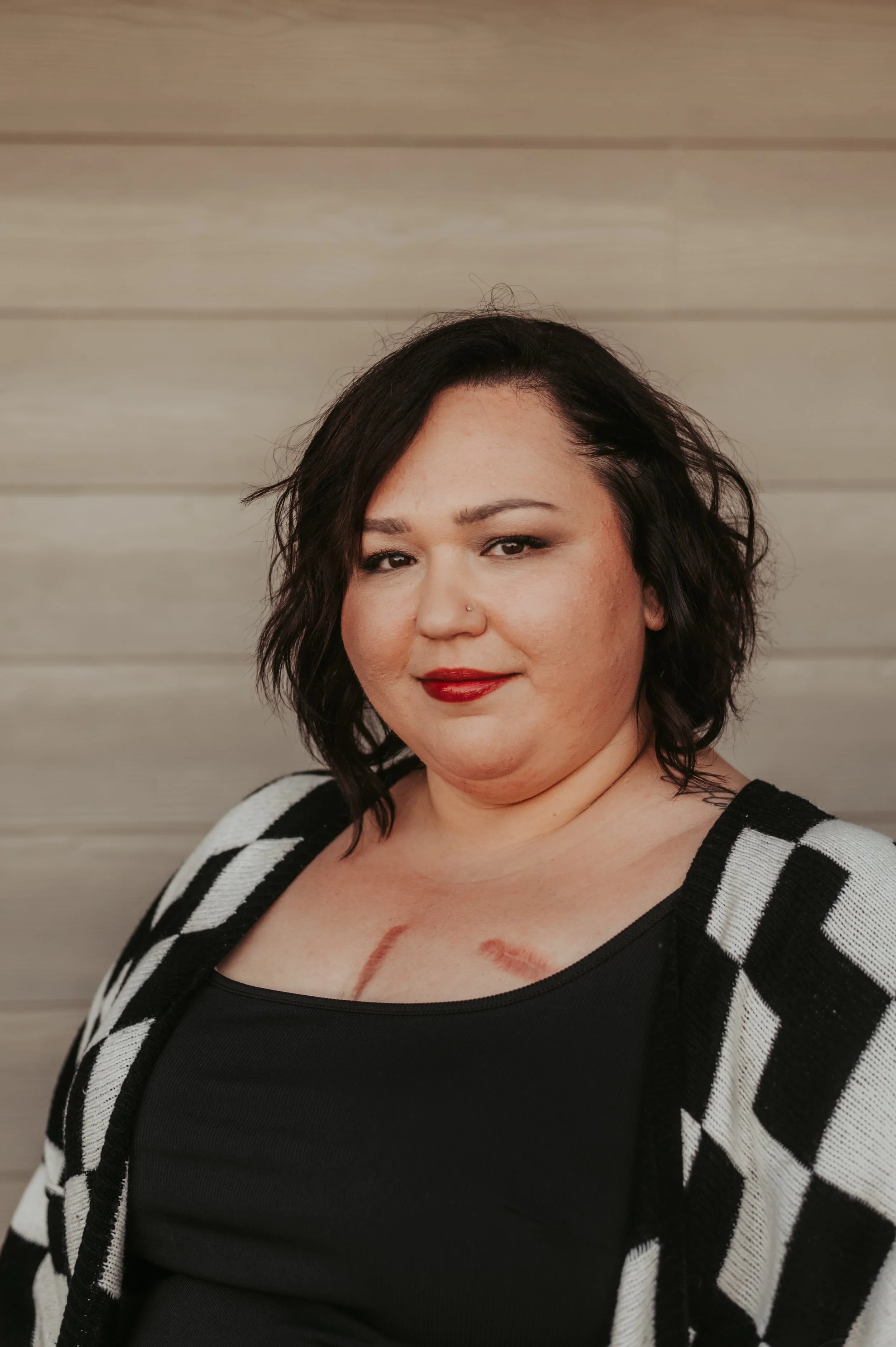 A woman with shoulder-length dark hair, wearing a black top and a black and white checkered cardigan, standing against a light wooden wall, with visible lipstick marks on her chest.