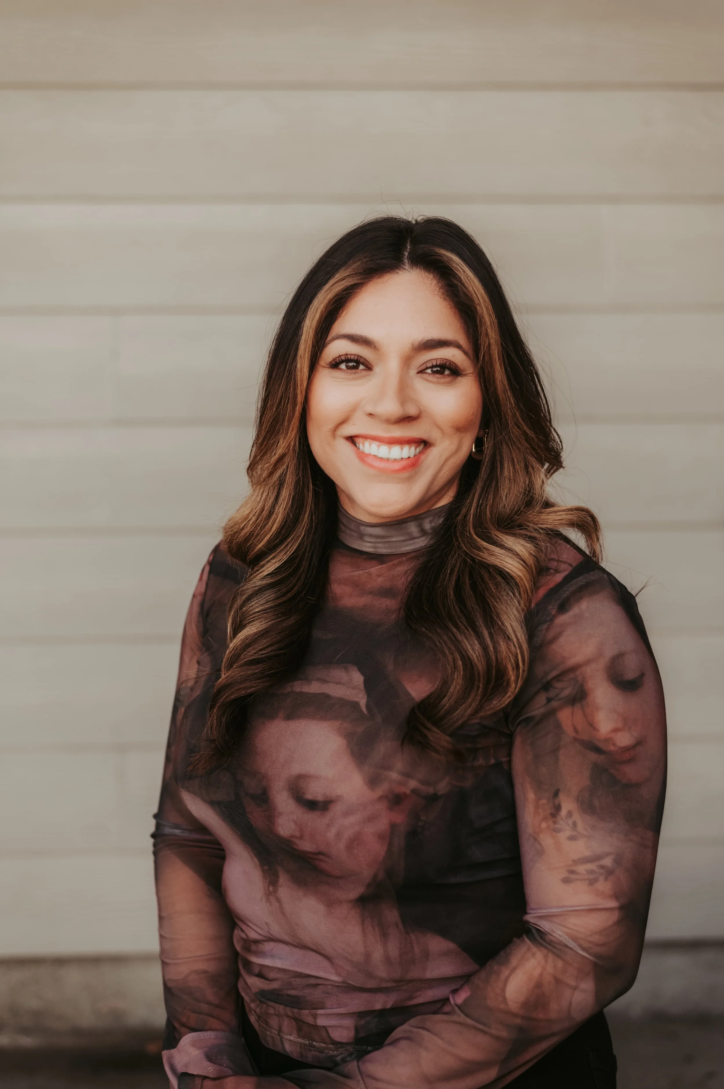 A woman with long, wavy brown hair smiling at the camera, wearing a sheer, dark-colored top with artistic printed images of faces on it, standing against a neutral background.