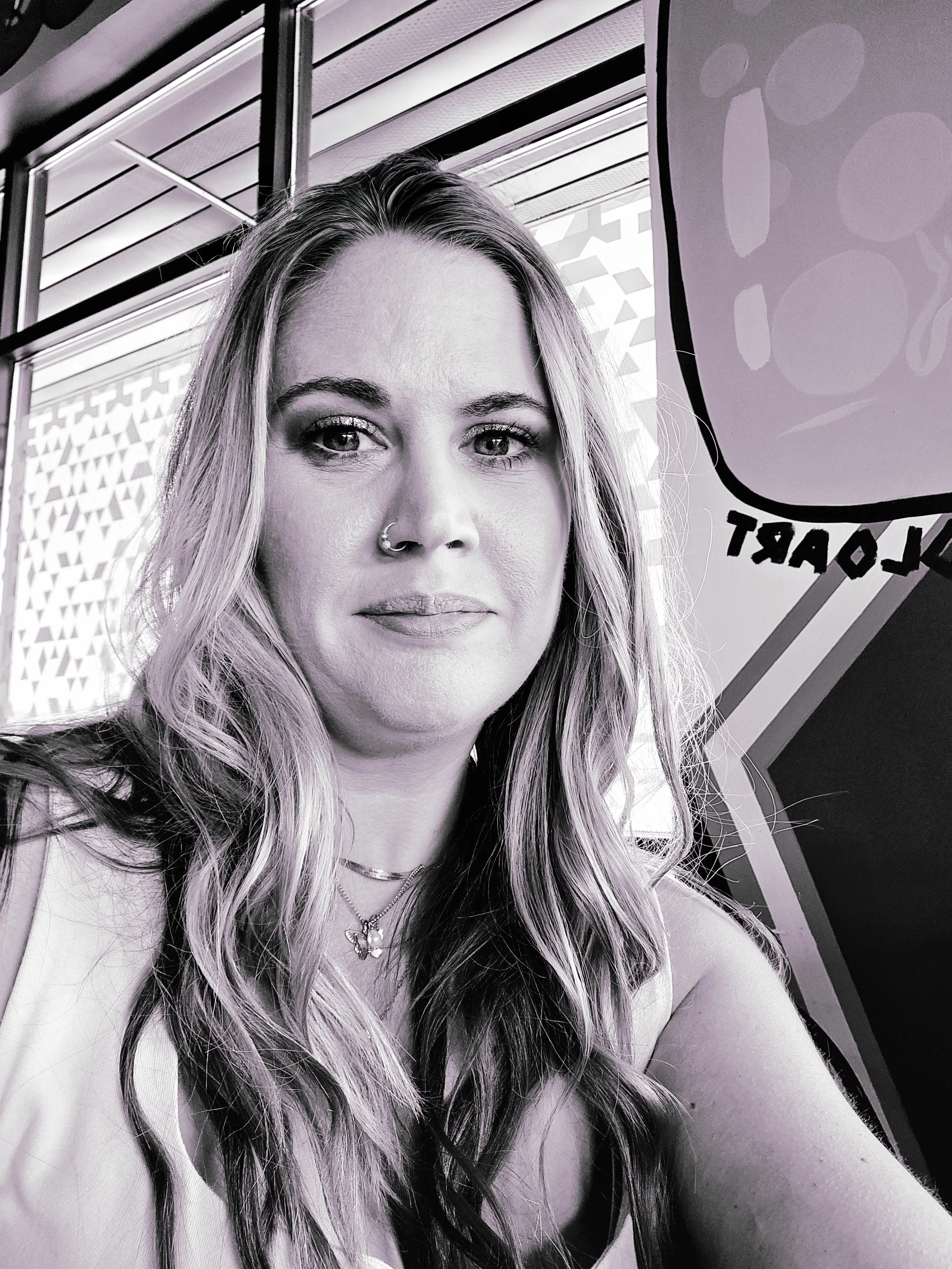 A woman with long wavy hair, wearing a nose ring, layered necklaces, and a sleeveless top, sitting indoors next to a window with striped blinds and a colorful wall with abstract art.