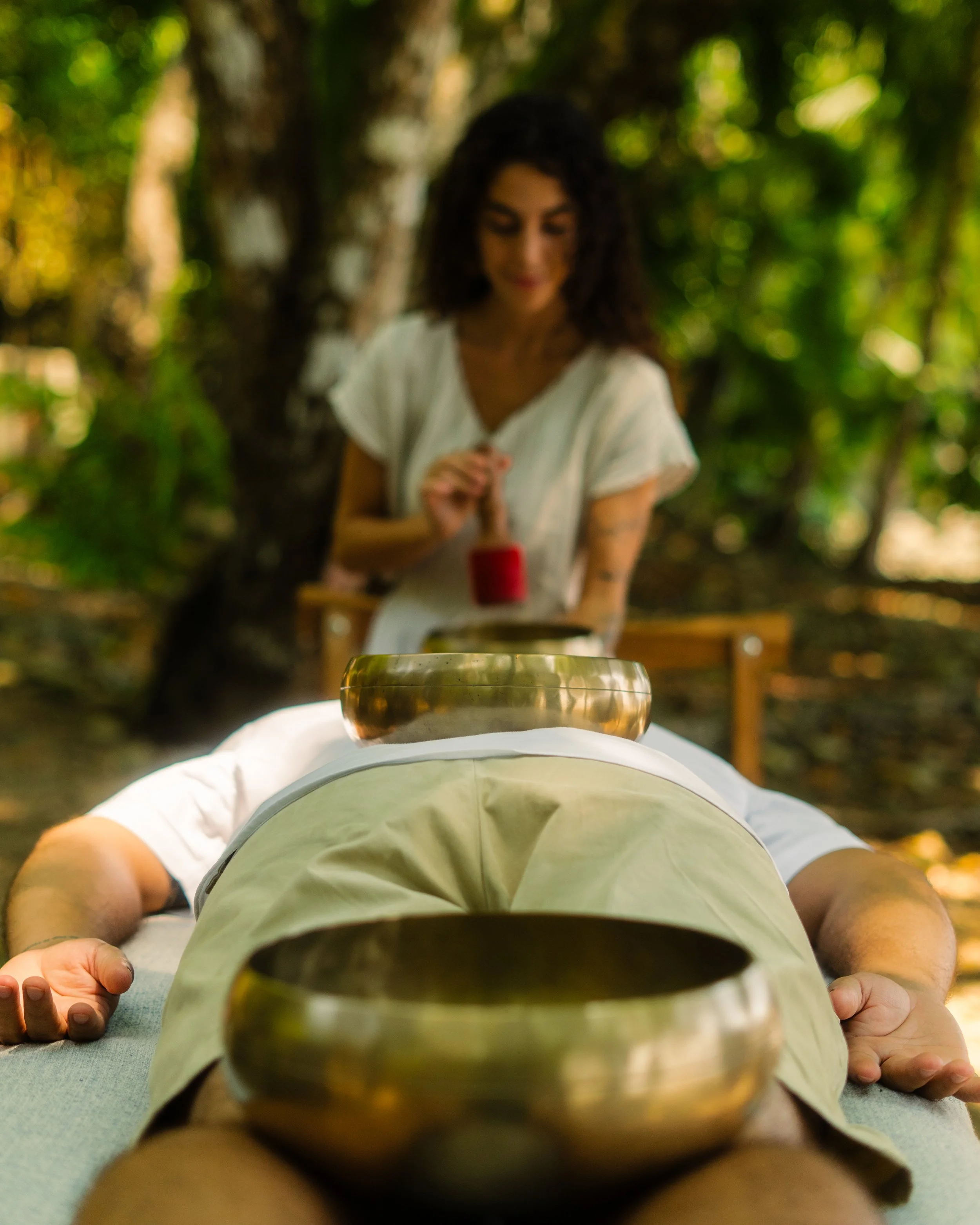 Woman playing a tibetan bowl over a man laying and relaxed, outdoors on the beach.