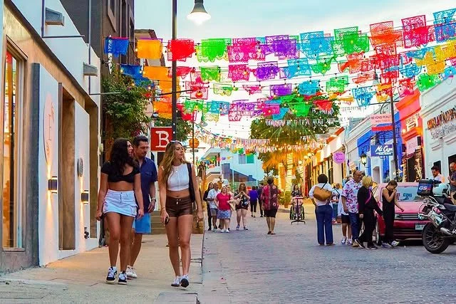 Street decorated with colorful hanging paper banners, people walking and shopping, buildings with shops and cafes, a palm tree, and a clear sky.