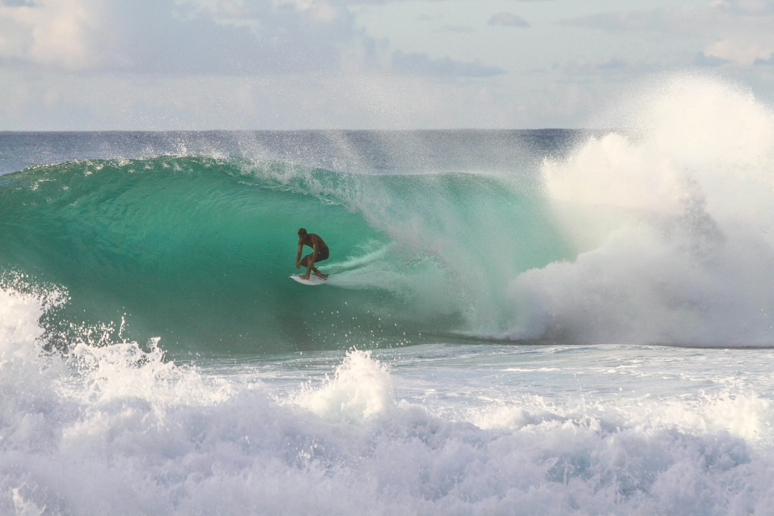 A man surfing inside a large turquoise wave near the shoreline, with spray and foam around him under a cloudy sky.