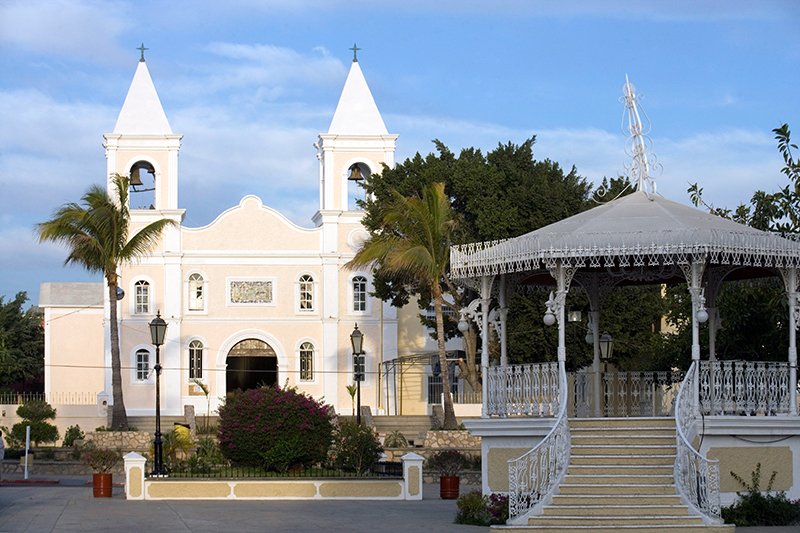 A white church with twin steeples and a small ornate white gazebo in front, surrounded by palm trees and greenery, under a partly cloudy sky.