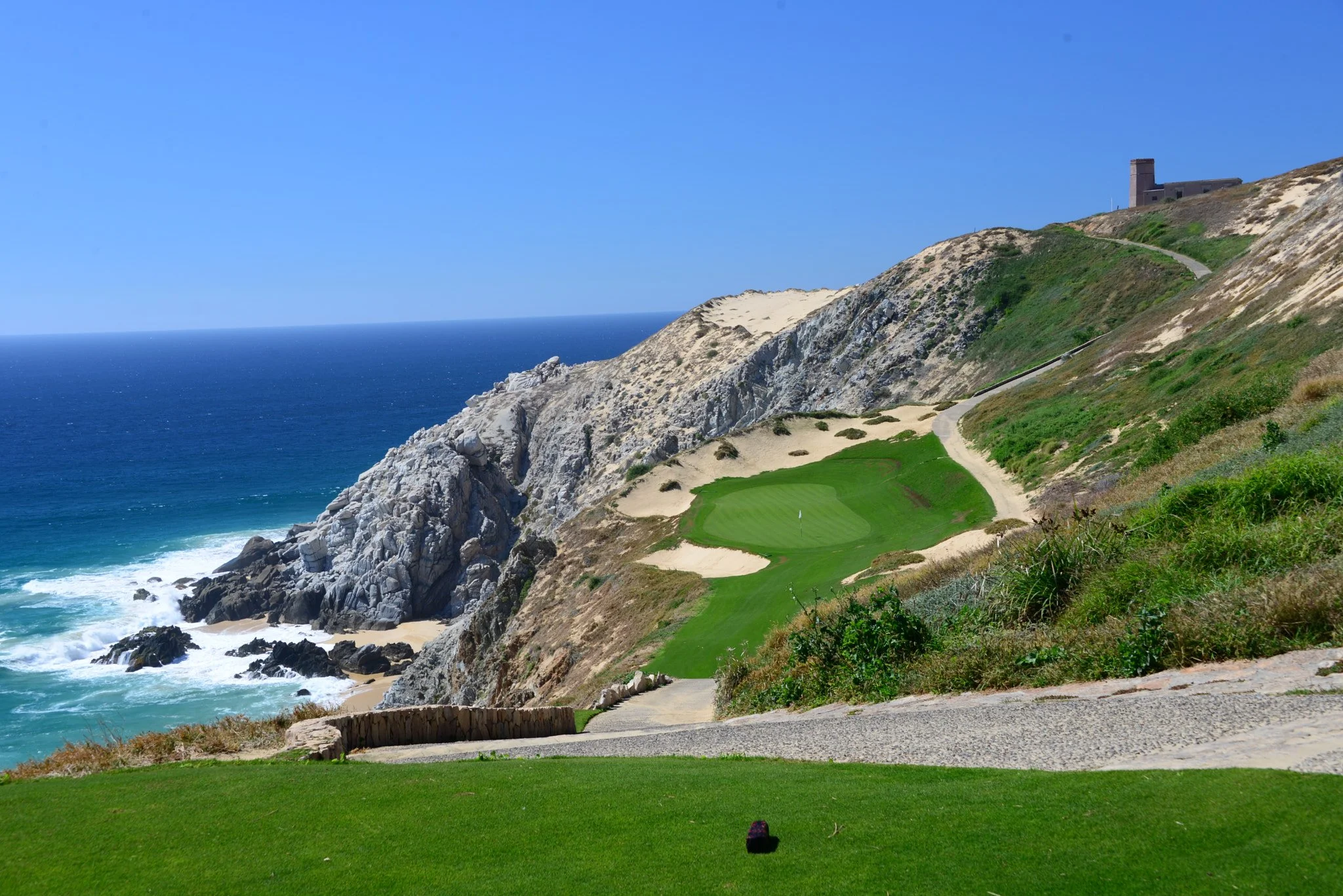 A golf course on a cliffside overlooking the ocean with a sandy beach, rocky shoreline, and a building on the hilltop under a blue sky.