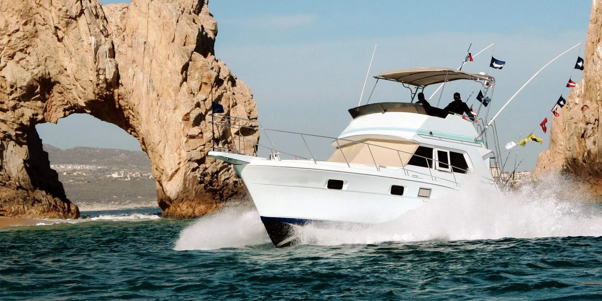 A white motor yacht with a canopy, flying multiple nautical flags, speeding through water near rocky cliffs with an arch formation.