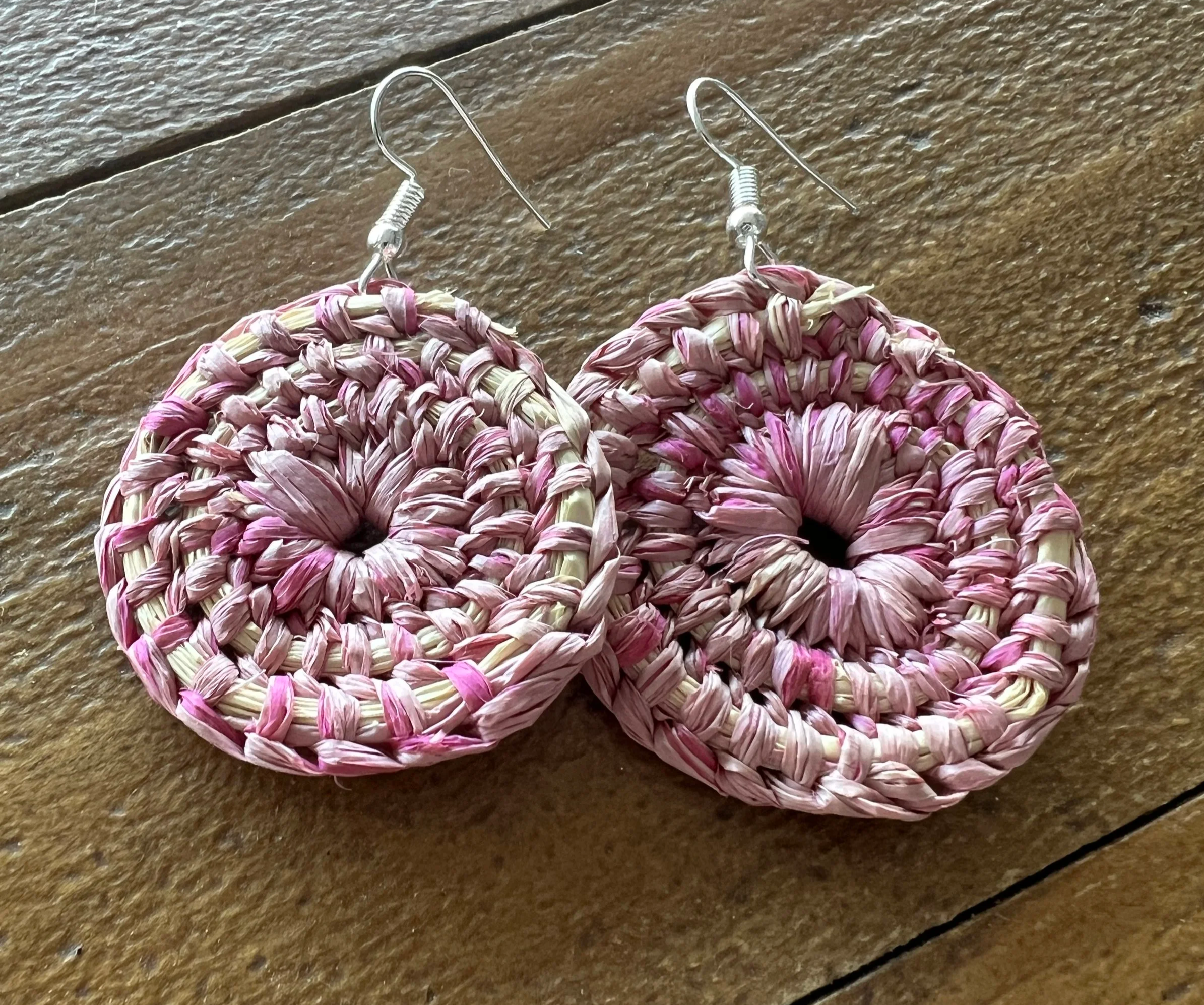 Circular woven pink and beige earrings with metal hooks, lying on a wooden surface.