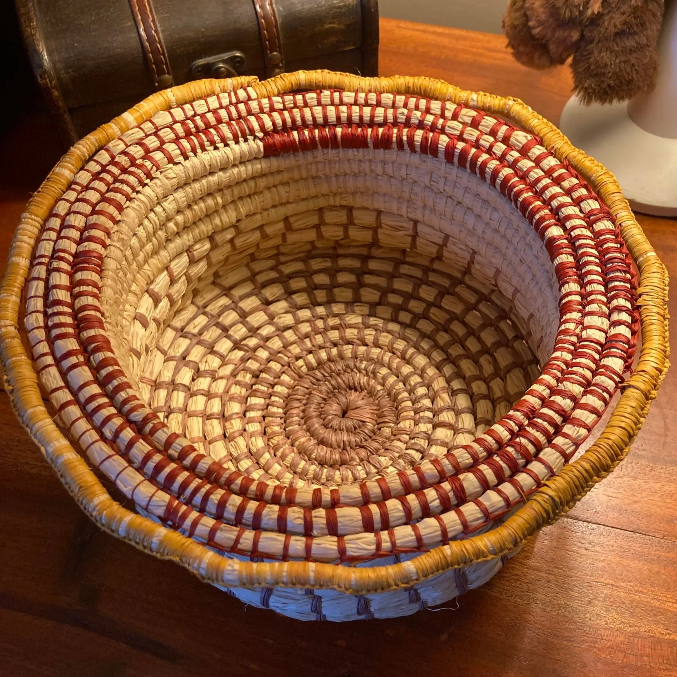 A circular woven basket containing natural raffia along with red, yellow and brown raffia.