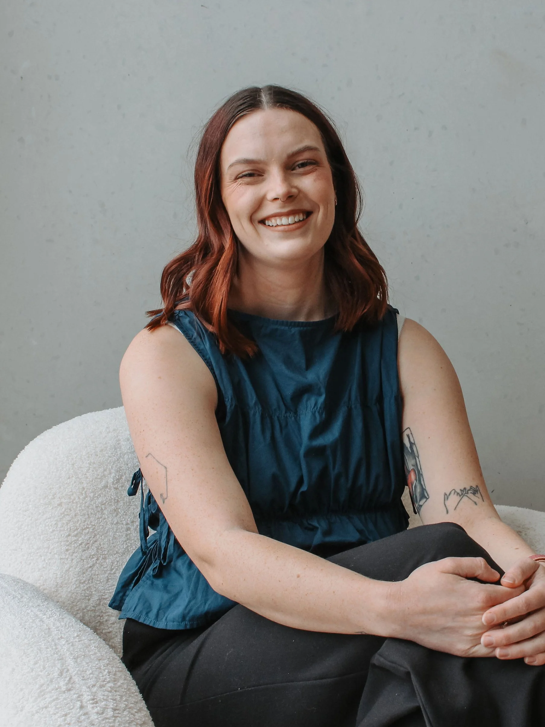 A woman with shoulder-length red hair, smiling, wearing a sleeveless blue top and black pants, sitting on a white textured cushion against a plain light gray wall, with visible tattoos on her arms.