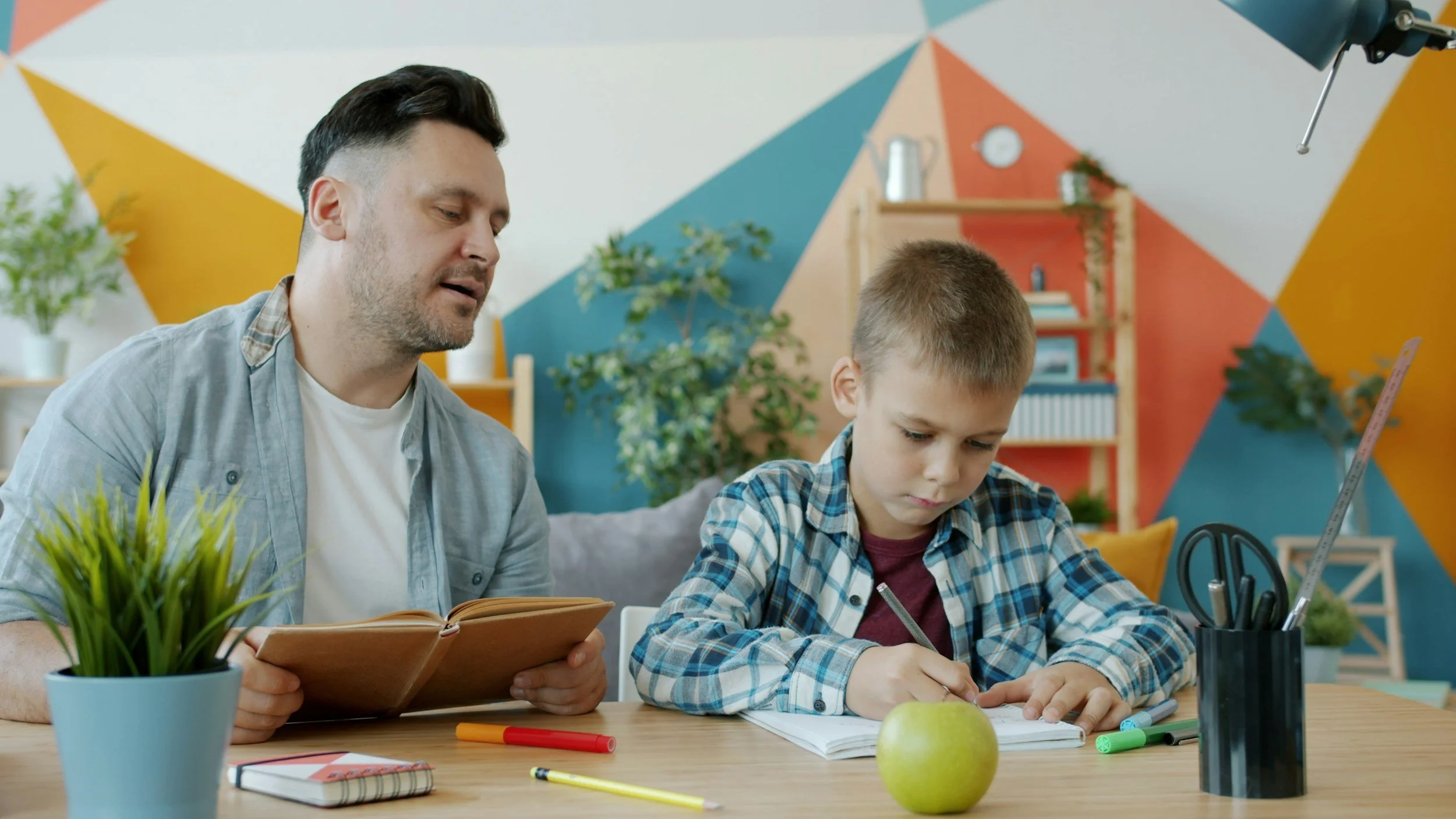 A man and a boy sitting at a table, reading a book and writing, with school supplies and a green apple on the table, in a colorful room with plants and geometric wall art.