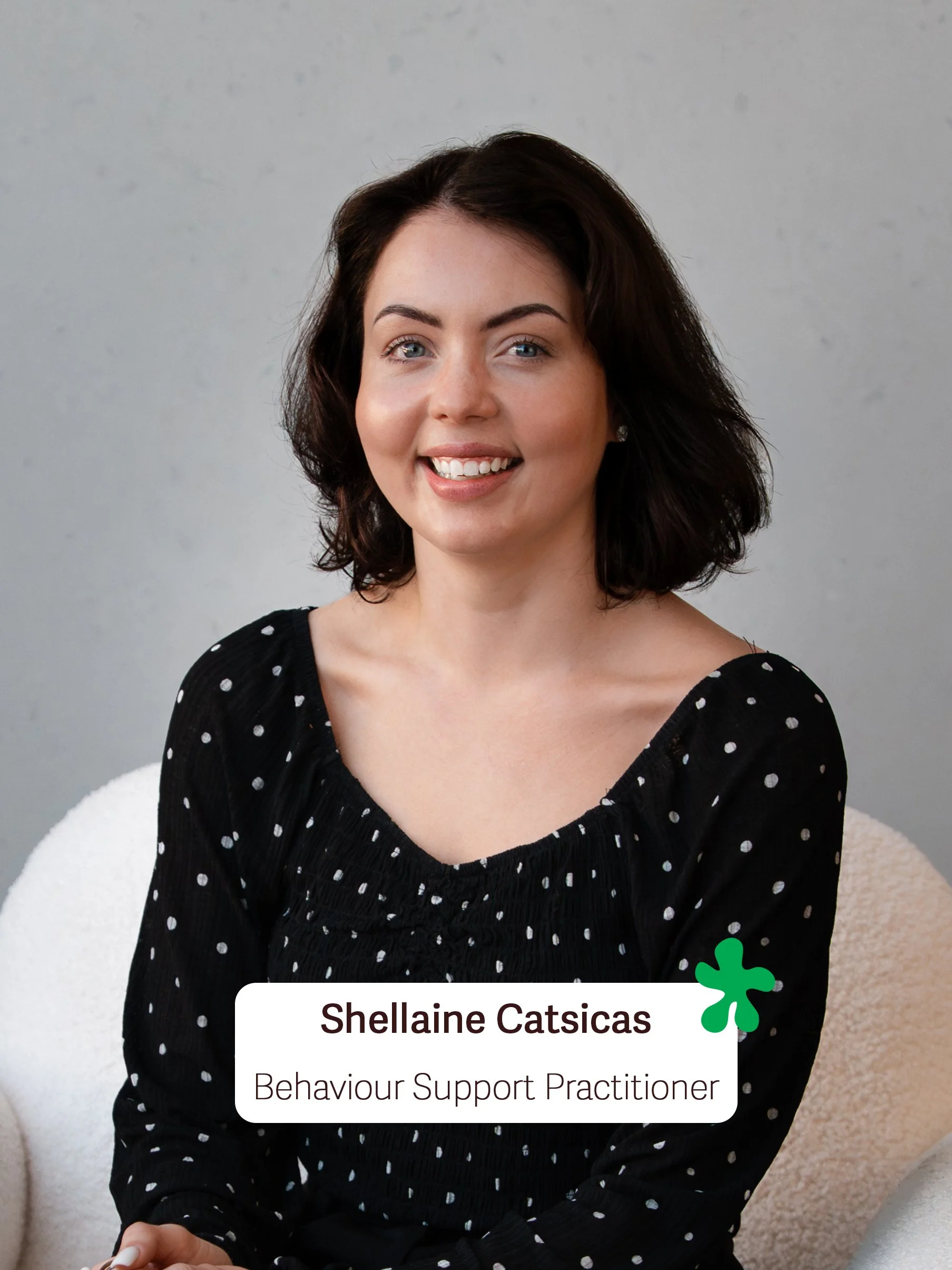 A woman with dark wavy hair, light skin, and blue eyes smiling while sitting in front of a plain light gray wall. She is wearing a black top with white polka dots. A label on her chest reads 'Shellaine Catsicas, Behaviour Support Practitioner' with a green decorative element next to her name.