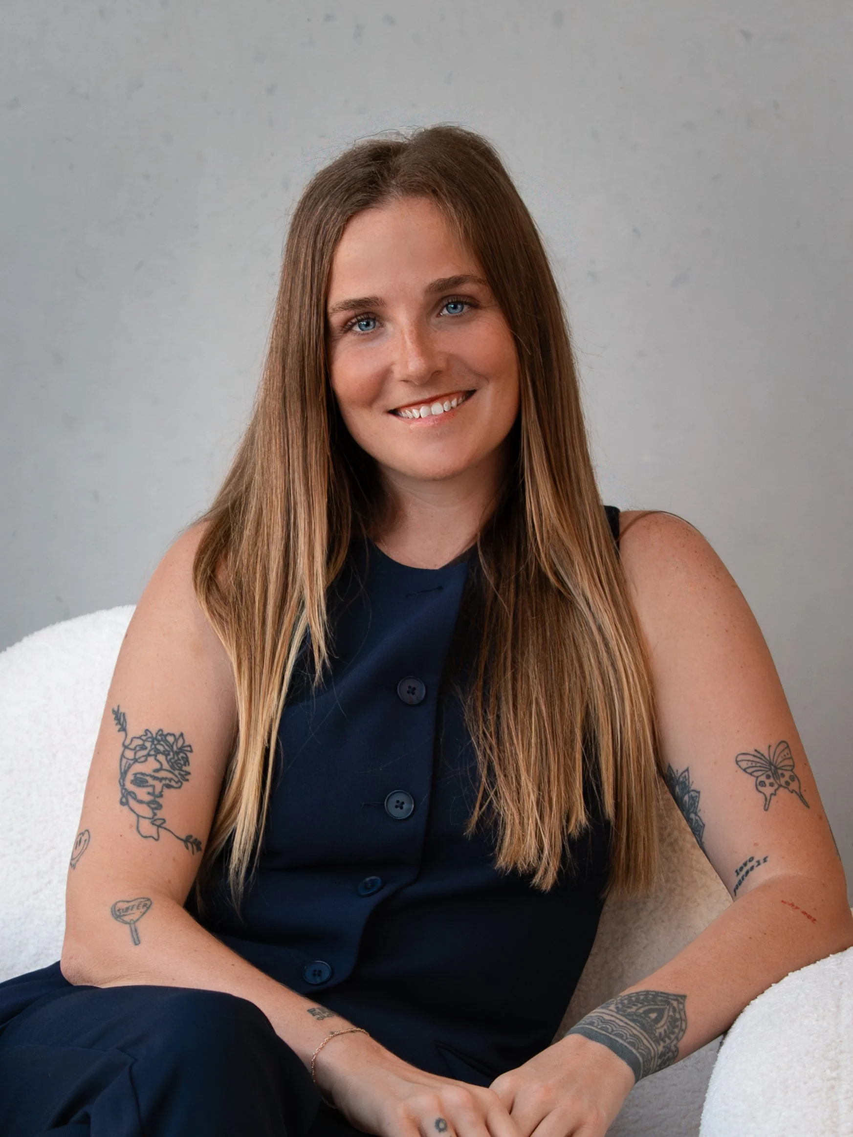 A woman with long brown hair and blue eyes smiling, sitting on a white towel against a plain light gray wall. She is wearing a dark sleeveless dress with large buttons and has visible tattoos on her arms.