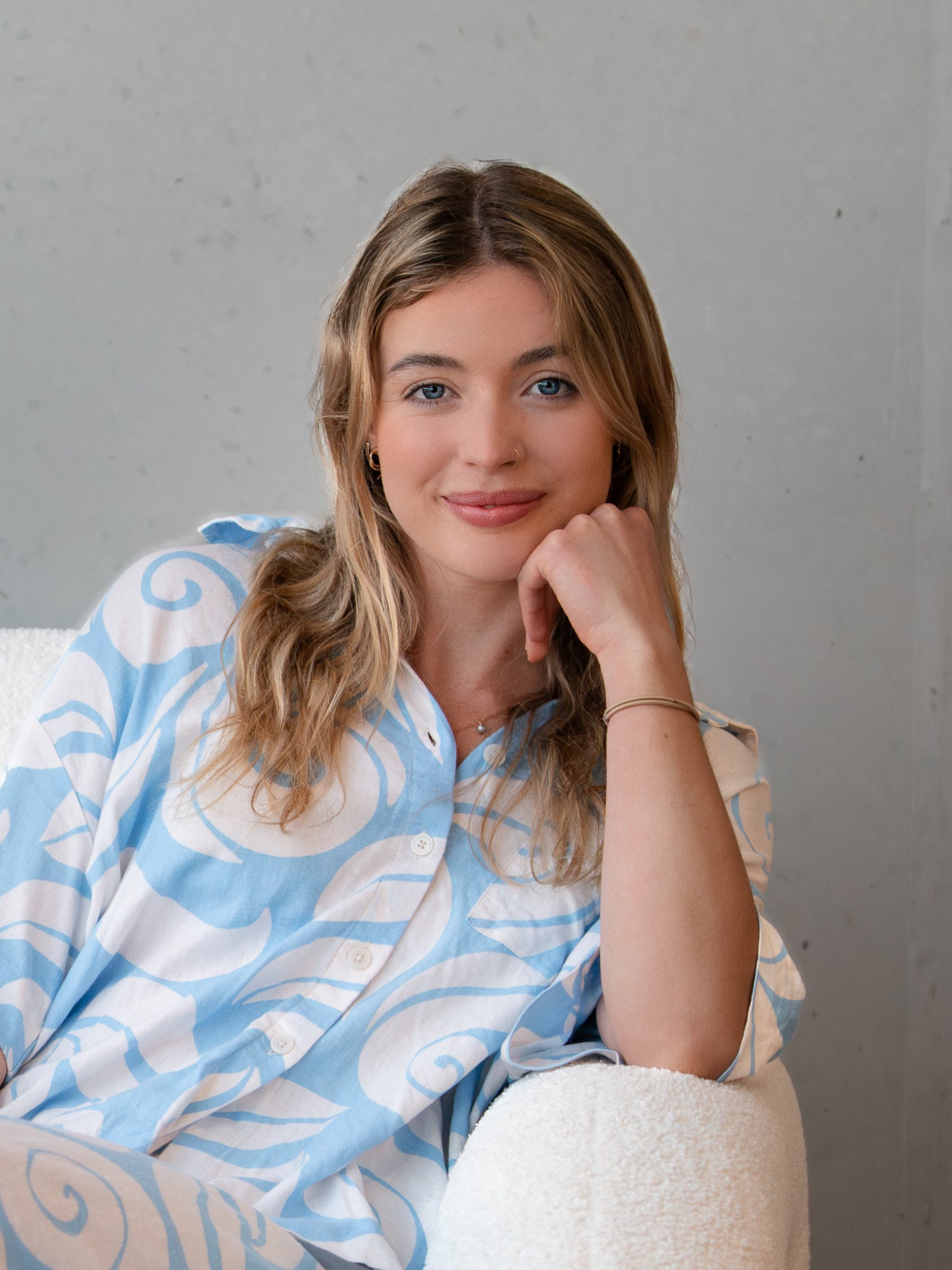 A young woman with blonde wavy hair and blue eyes sitting indoors against a neutral wall, resting her chin on her hand, wearing a light blue and white patterned button-up shirt.