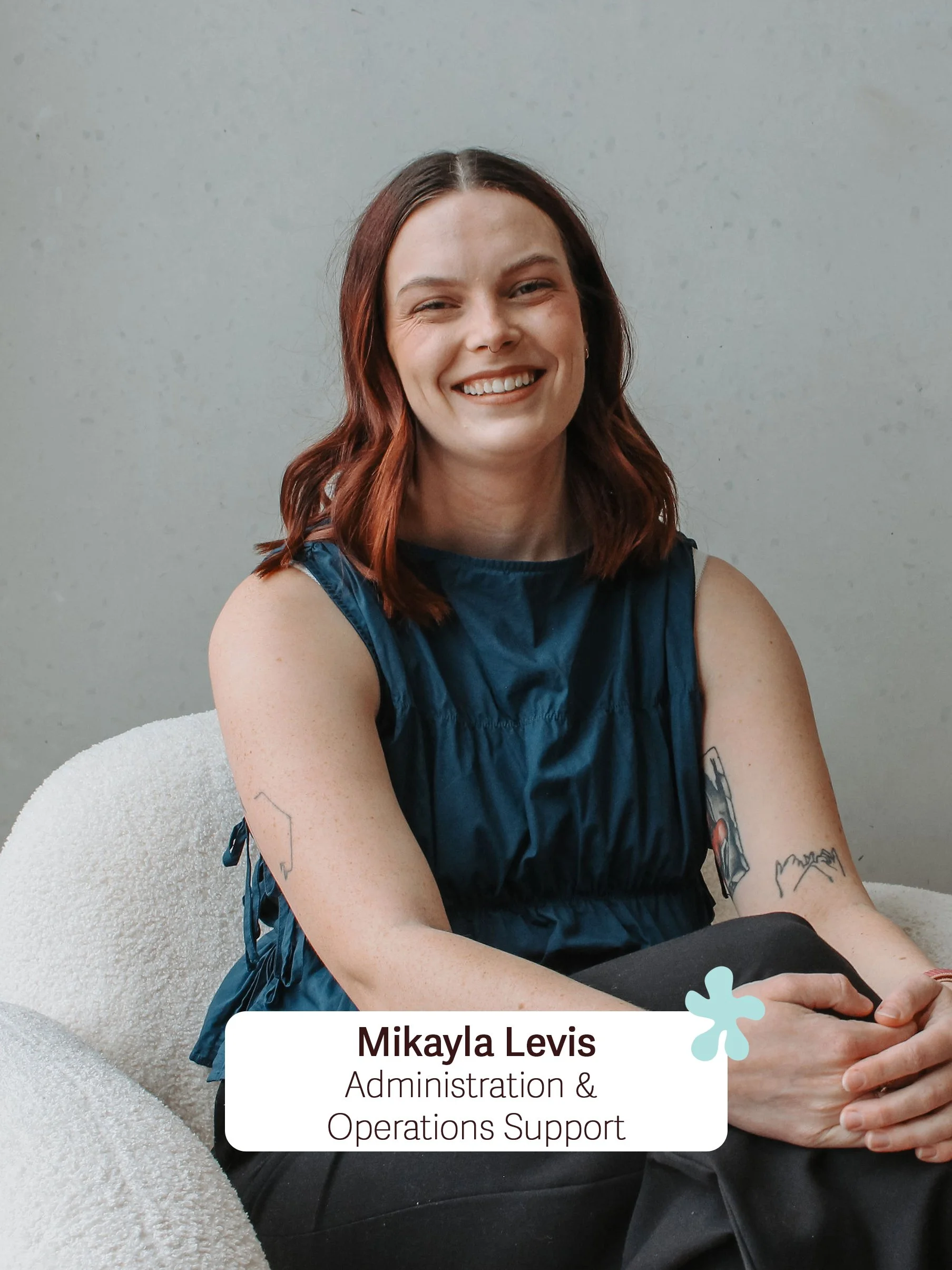 A woman with shoulder-length red hair, smiling, sitting on a white textured chair against a plain light gray wall. She is wearing a sleeveless dark blue top and has tattoos on her left arm.