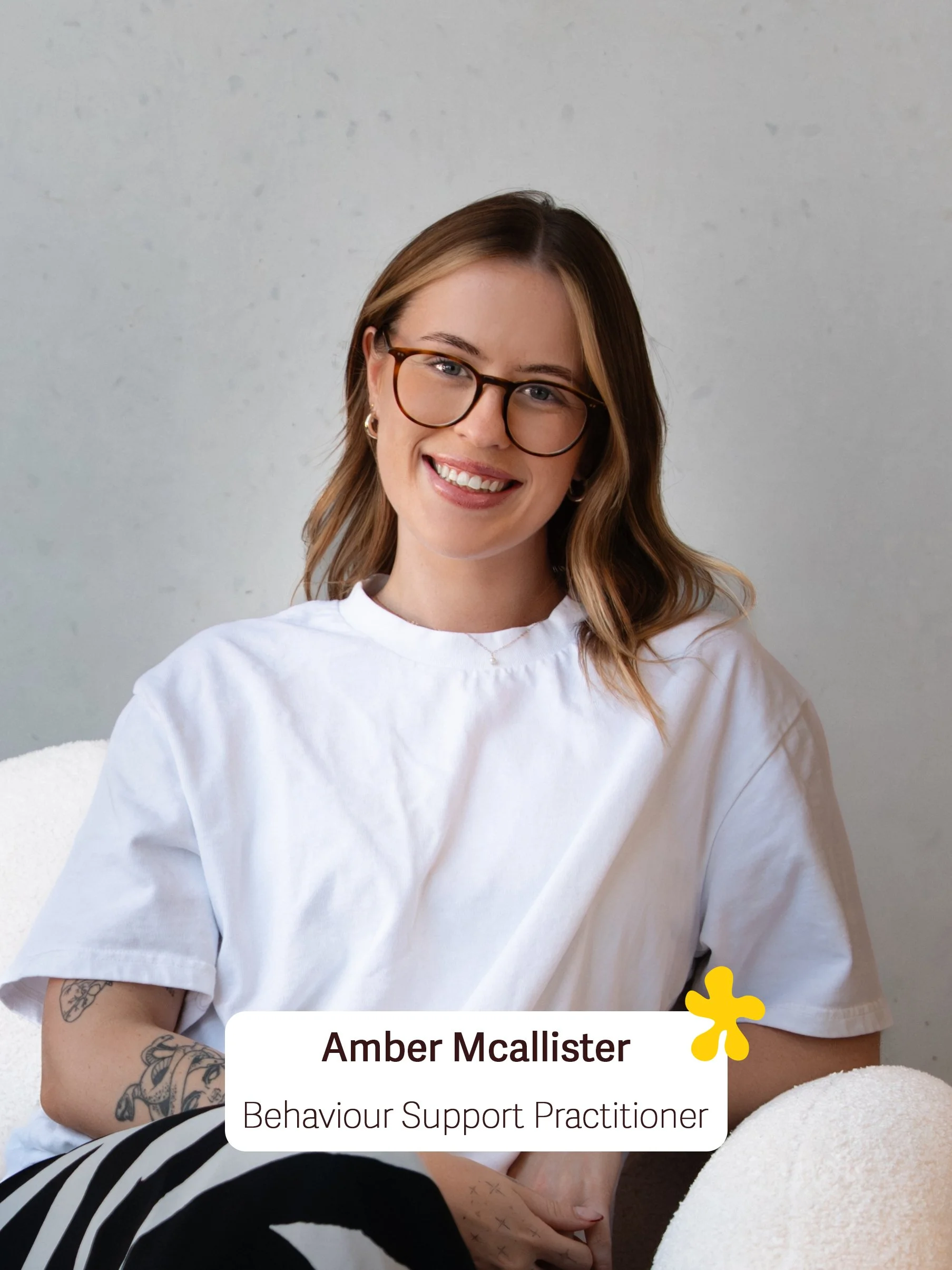 A woman with shoulder-length brown hair, glasses, and a white T-shirt, sitting on a white textured surface with a neutral background. She is smiling and has tattoos on her left arm. Overlaid text reads: "Amber Mcallister, Behaviour Support Practitioner".