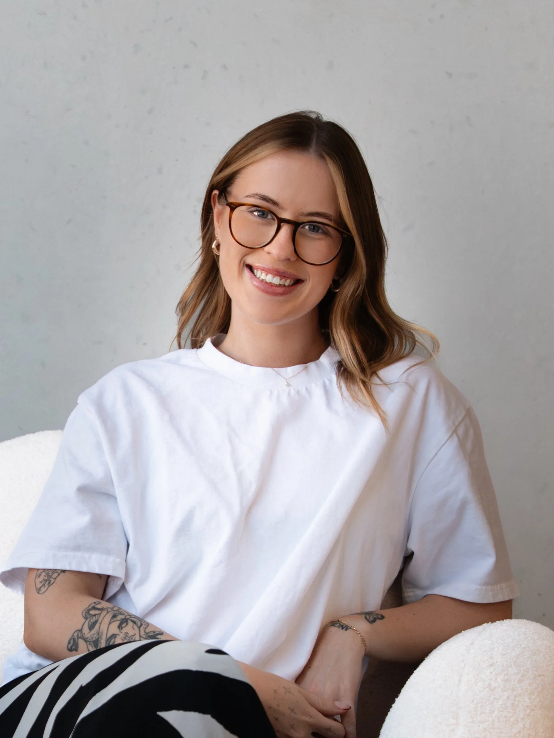 A woman with shoulder-length brown hair, wearing glasses and a white T-shirt, smiling while sitting on a white chair with a white cushioned armrest, in front of a plain gray wall.