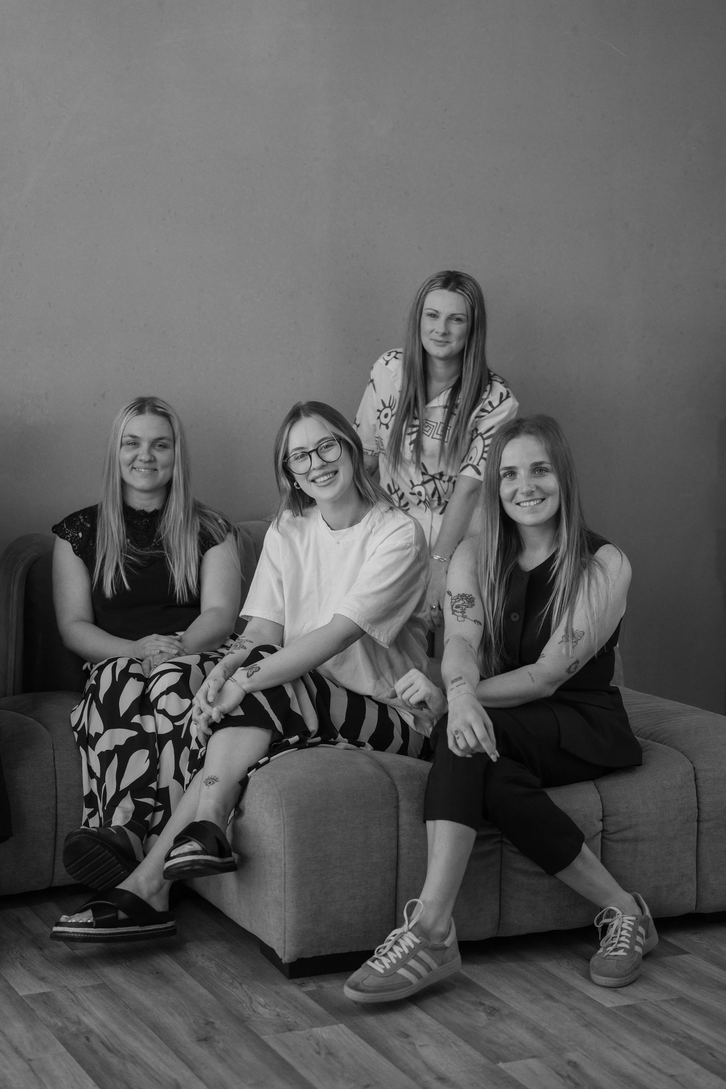 Four young women sitting and standing on a sofa in a room, smiling and posing for the photo, with a plain wall in the background.