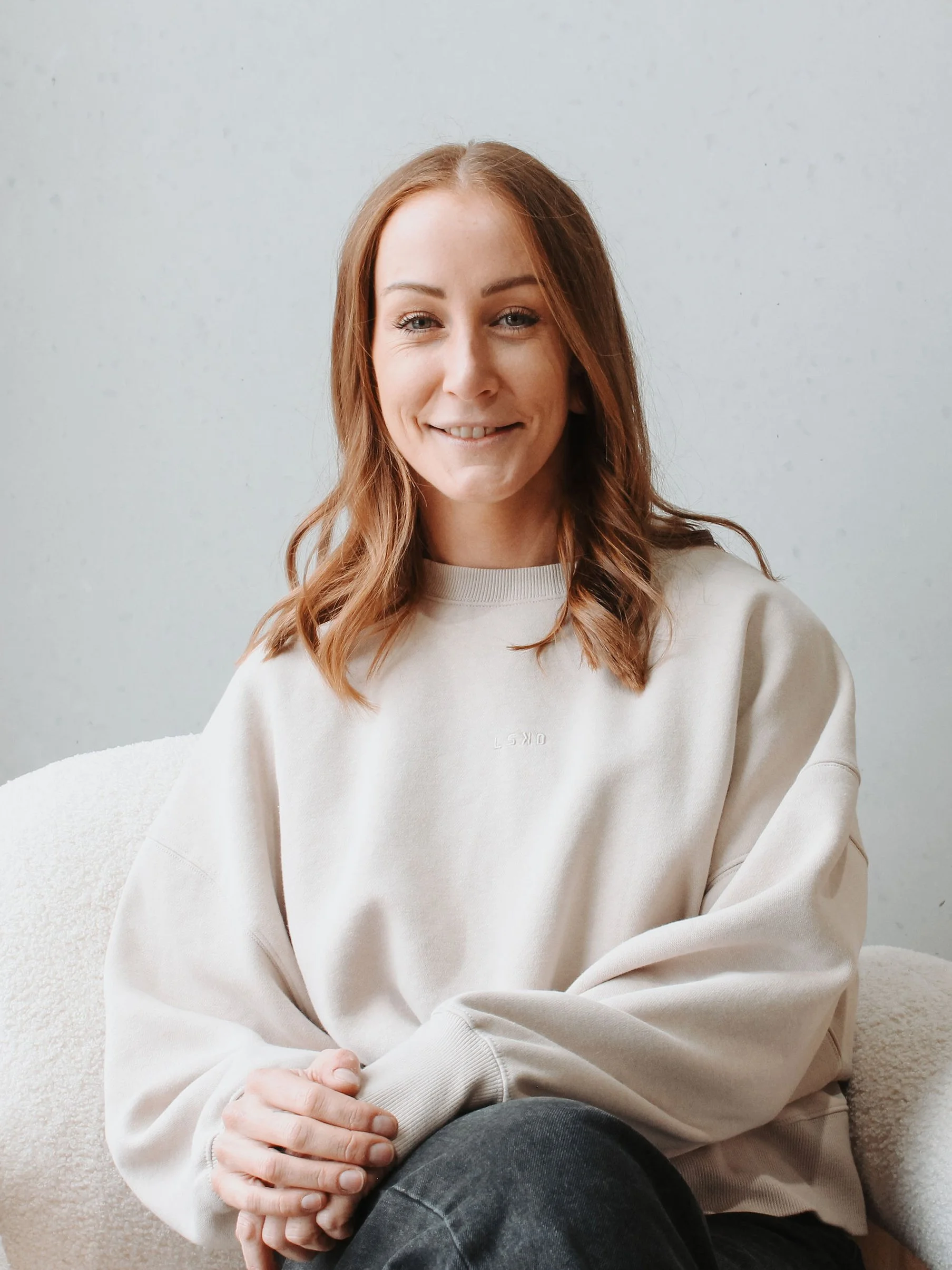 A young woman with light skin and red hair sitting on a beige couch, wearing a cream-colored sweatshirt and dark jeans, smiling at the camera with one hand resting on her knee.