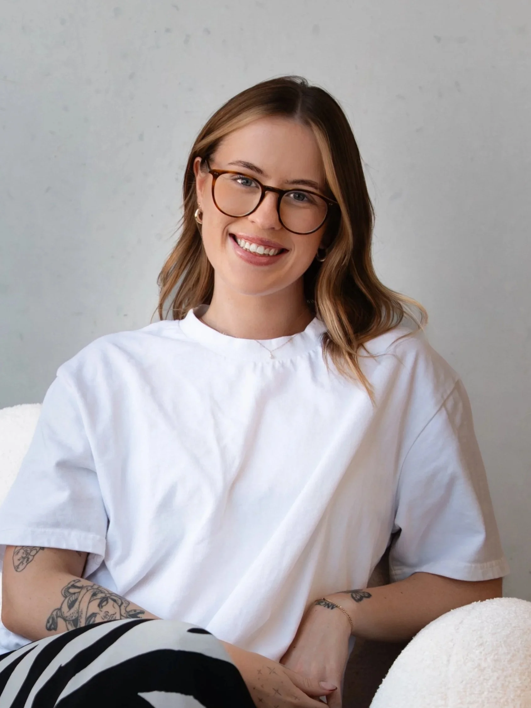 A woman with light skin, brown hair with blonde highlights, wearing glasses and a white t-shirt, is smiling at the camera. She has tattoos on her forearm and wrist, and is sitting on a white surface against a gray wall background.