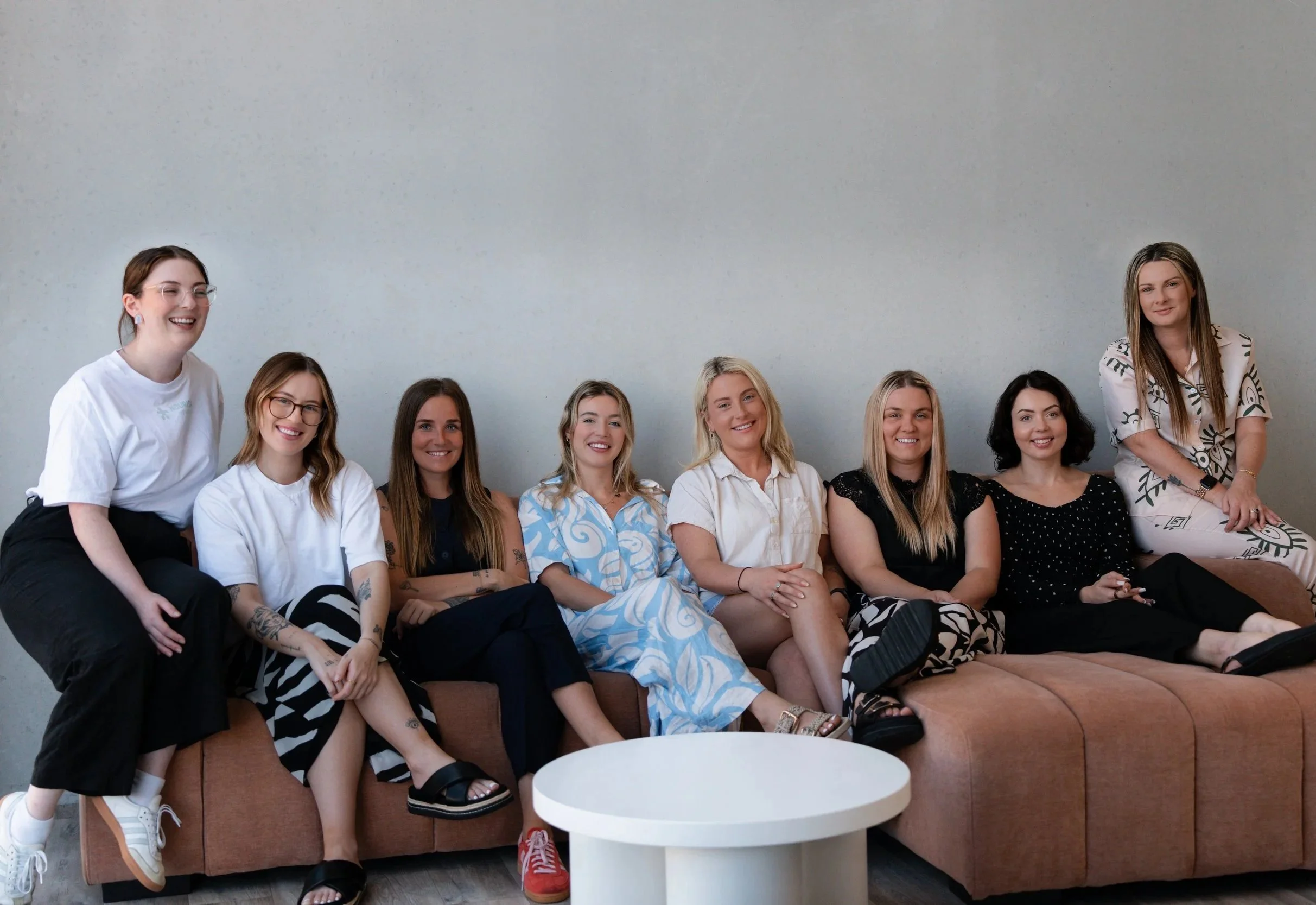 Ten women sitting on a brown sofa in front of a plain gray wall, smiling and posing for the photo.