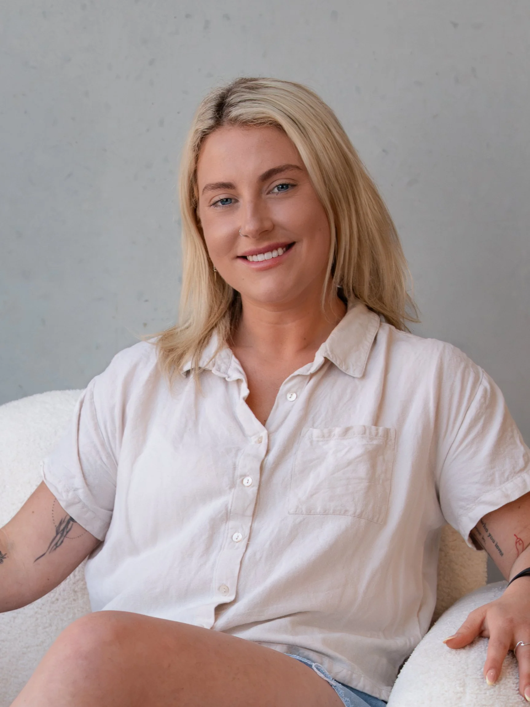 A woman with blonde hair, blue eyes, and a small nose, smiling, sitting on a beige couch, wearing a beige button-up shirt, with tattoos on her left arm, in a minimalistic room with a plain background.