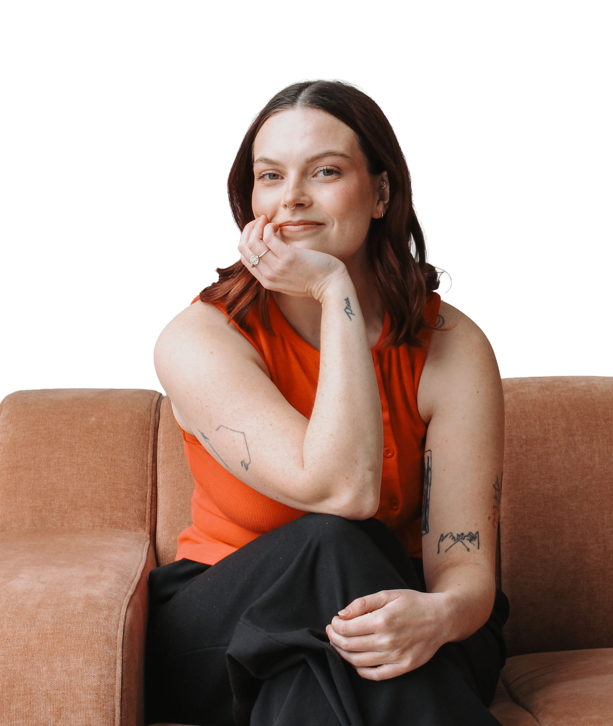 A woman with reddish-brown hair and multiple tattoos, wearing an orange sleeveless top and black pants, sitting on a tan sofa, resting her chin on her hand, looking at the camera with a gentle smile.