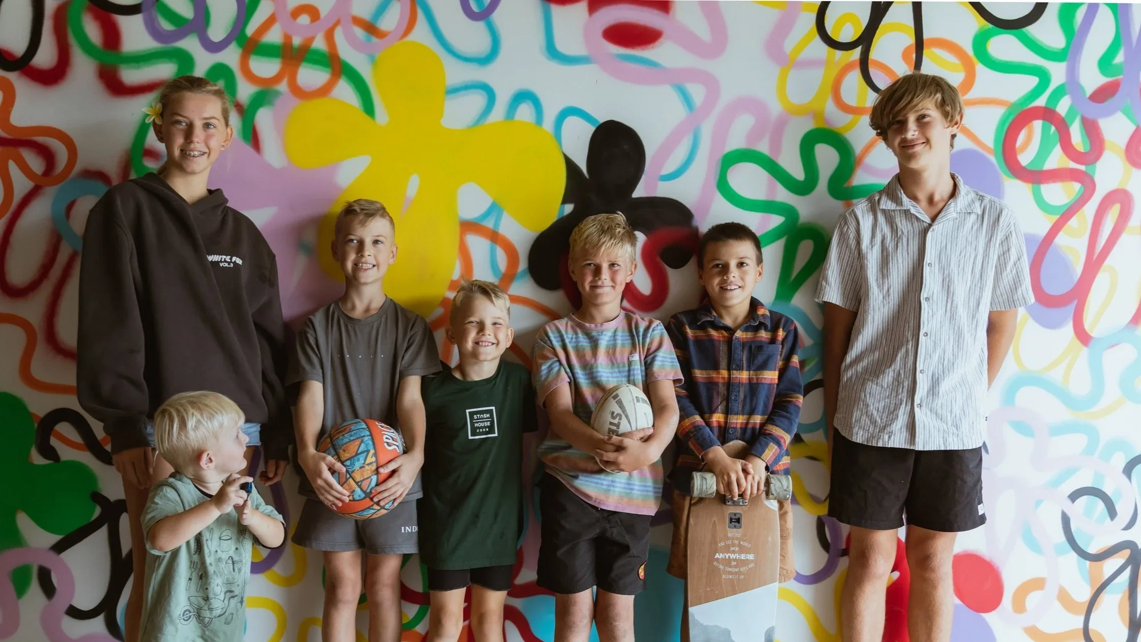 Group of children and teenagers, some holding skateboards and basketballs, standing in front of a colorful graffiti wall with abstract shapes and bright colors.