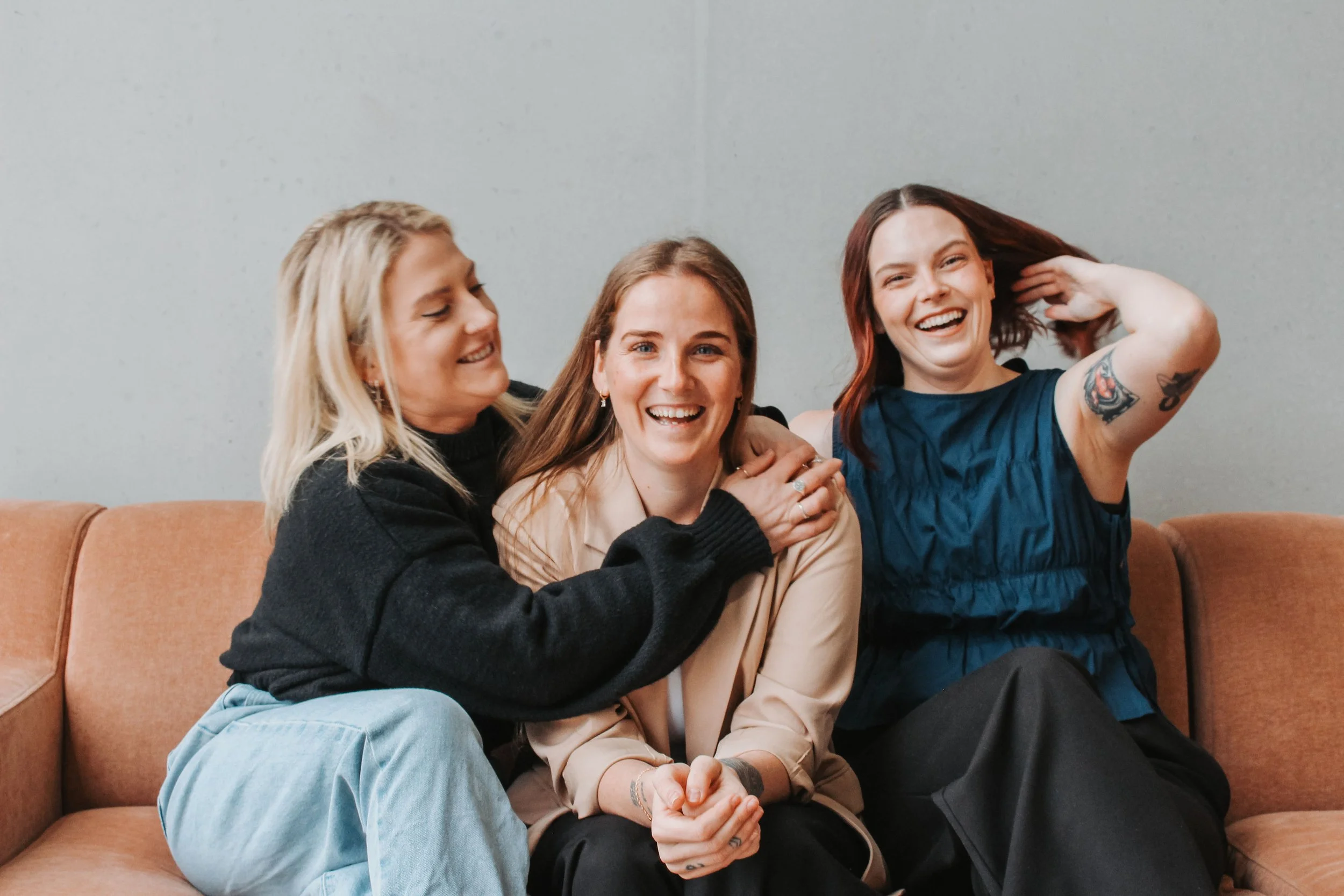 Three women sitting on a brown couch against a plain wall, smiling and laughing. The woman on the right has red hair and tattoos on her arm, and is holding her hair. The woman in the middle has brown hair and is wearing a beige blazer. The woman on the left has blonde hair and is wearing a black sweater, leaning toward the others.