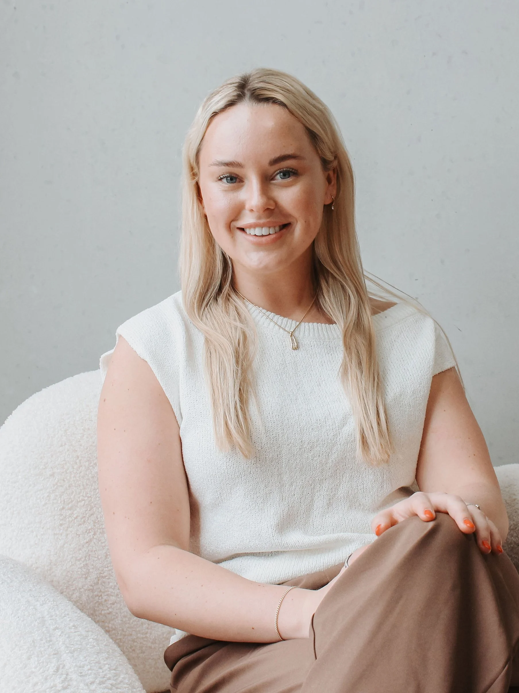 A young woman with blonde hair and blue eyes smiling while sitting on a cream-colored chair, wearing a white sleeveless top and beige pants, and jewelry including earrings, a necklace, a bracelet, and rings.