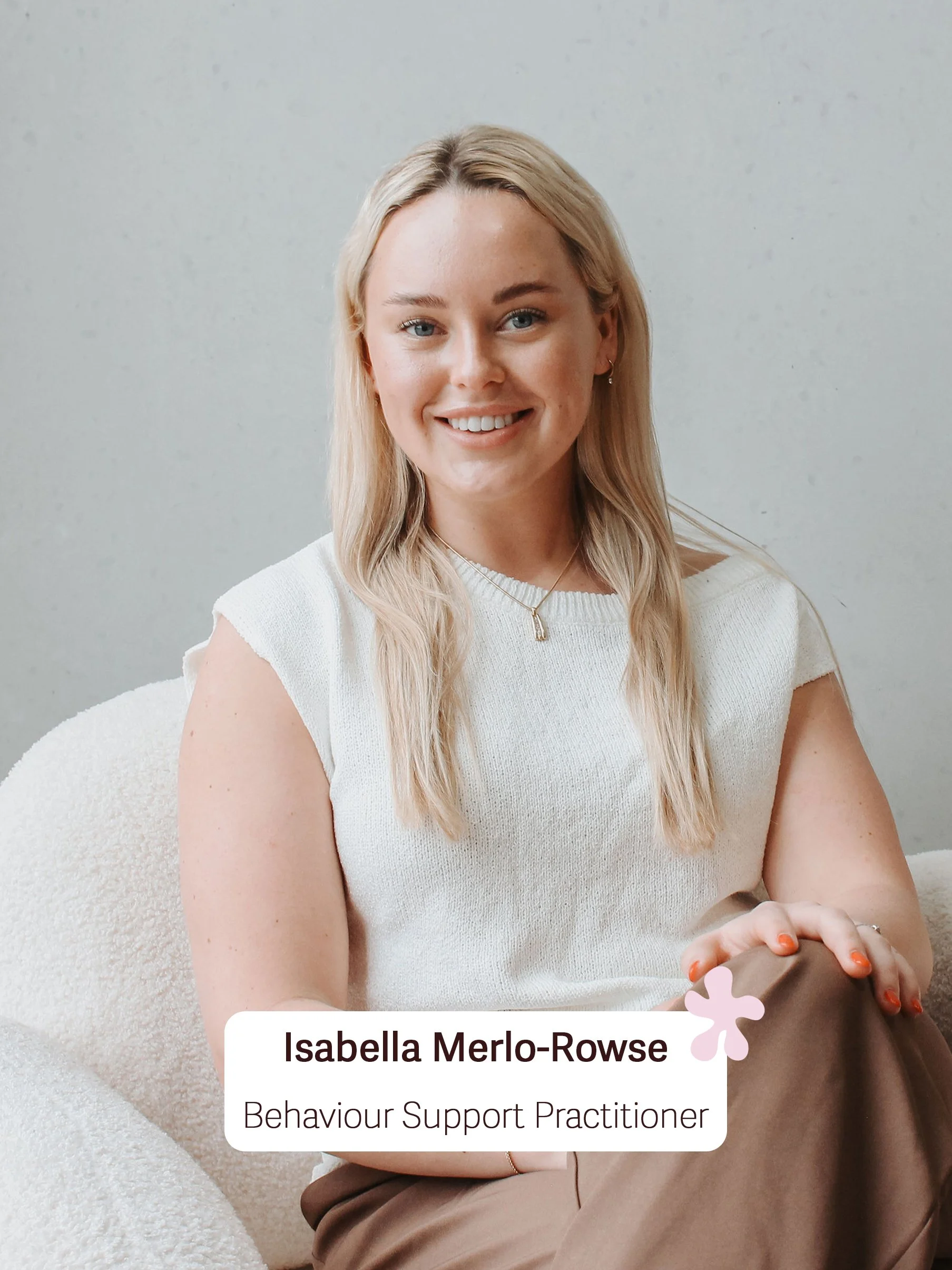 A woman with blonde hair smiling and wearing a white top, sitting on a light-colored plush chair, holding a small object in her hands. Text at the bottom reads: 'Isabella Merlo-Rowse, Behaviour Support Practitioner.'