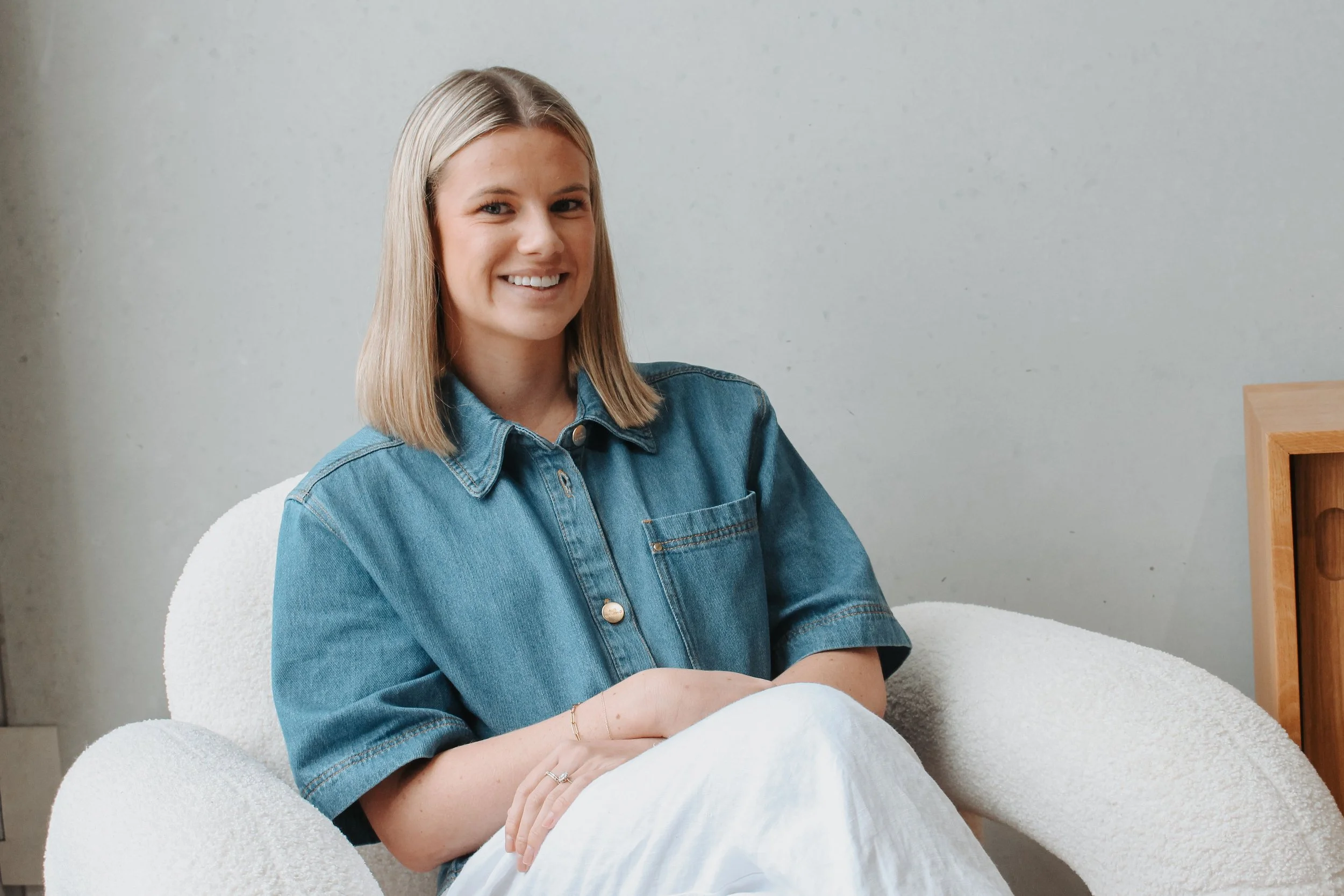 A woman with shoulder-length blonde hair, wearing a blue denim shirt, smiling, sitting on a white cushioned chair against a plain light-colored wall.