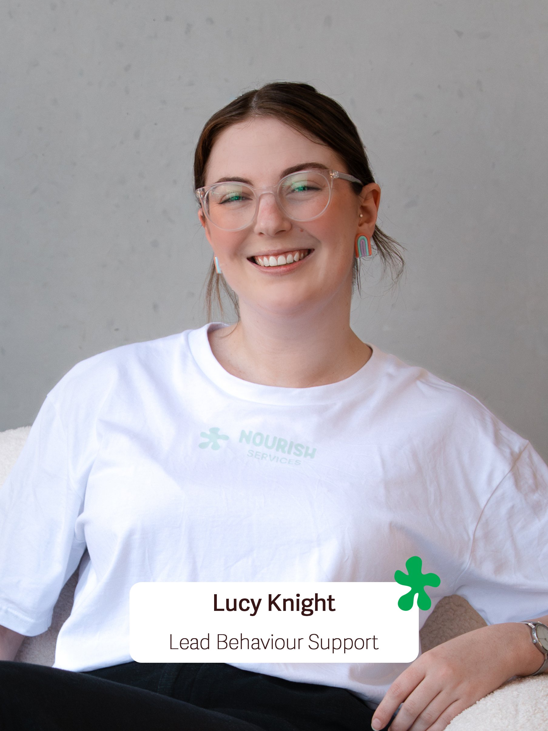 A young woman with short brown hair, glasses, and rainbow earrings, smiling and sitting on a light-colored surface against a plain gray background, wearing a white T-shirt with a green logo, holding a person's hand, with a watermarked text overlay that reads 'Lucy Knight, Lead Behaviour Support'.